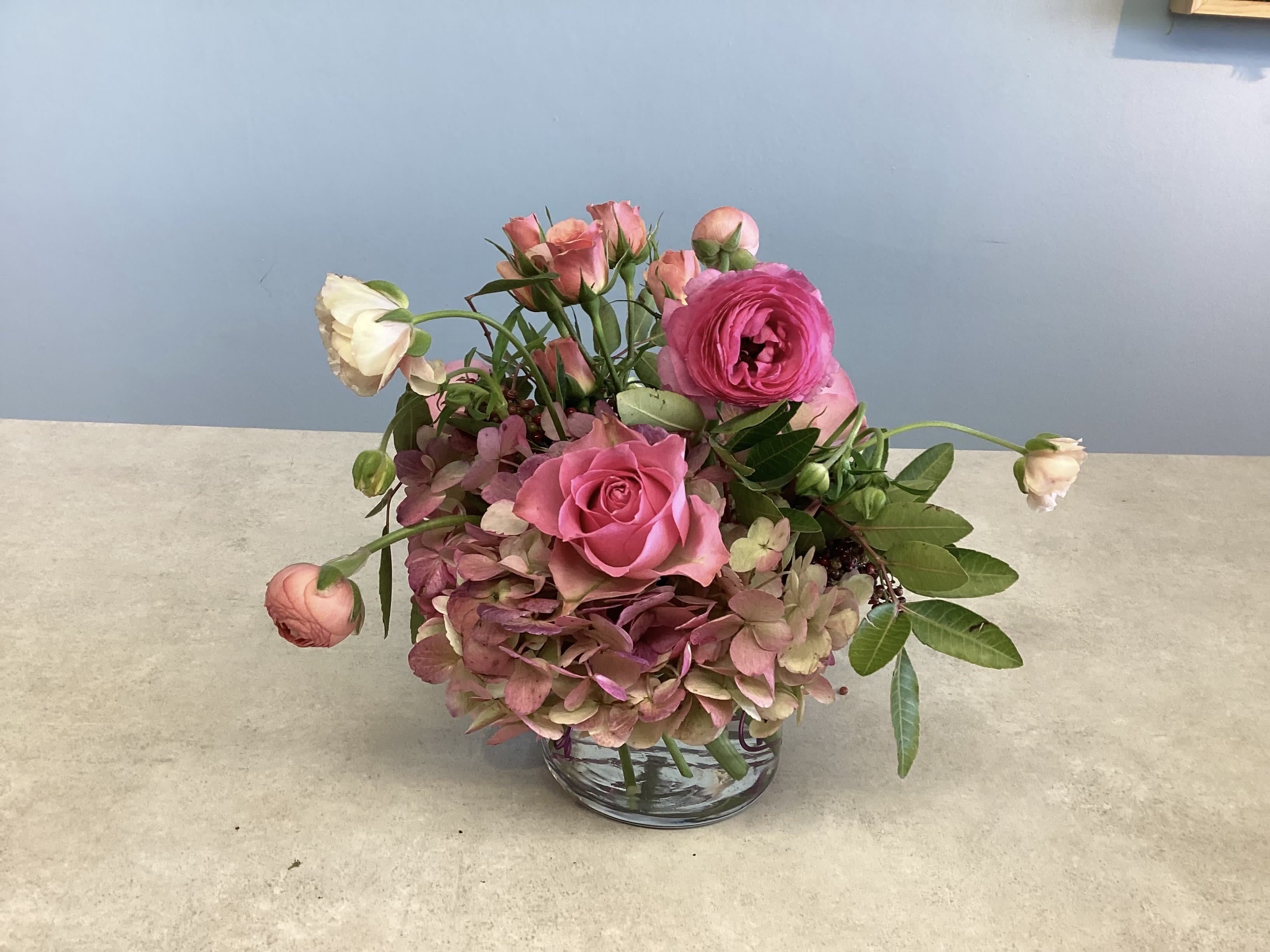 Meadow Muse - A compact garden-style arrangement in soft pinks, peaches and fresh green, Meadow Muse is a low, rounded flower arrangement perfect for a birthday bouquet, anniversary flowers, thank-you gift, or a just-because surprise. The mix of pink hydrangeas, roses and ranunculus, creates a romantic, feminine look that makes the person you're thinking of feel truly celebrated-intimate, fragrant, and effortlessly elegant.  Flowers are arranged tightly in a clear glass vase with a touch of decorative wire visible inside for a finished yet casual accent. Premium pink roses and lush pink hydrangea form the bulk of the composition, with pink ranunculus and filler blooms tucked around the edges and loose greenery framing the arrangement. Its low, rounded profile sits beautifully on a dining table or desk and arrives ready to display; light daily water checks will keep blooms looking fresh through their vase life.