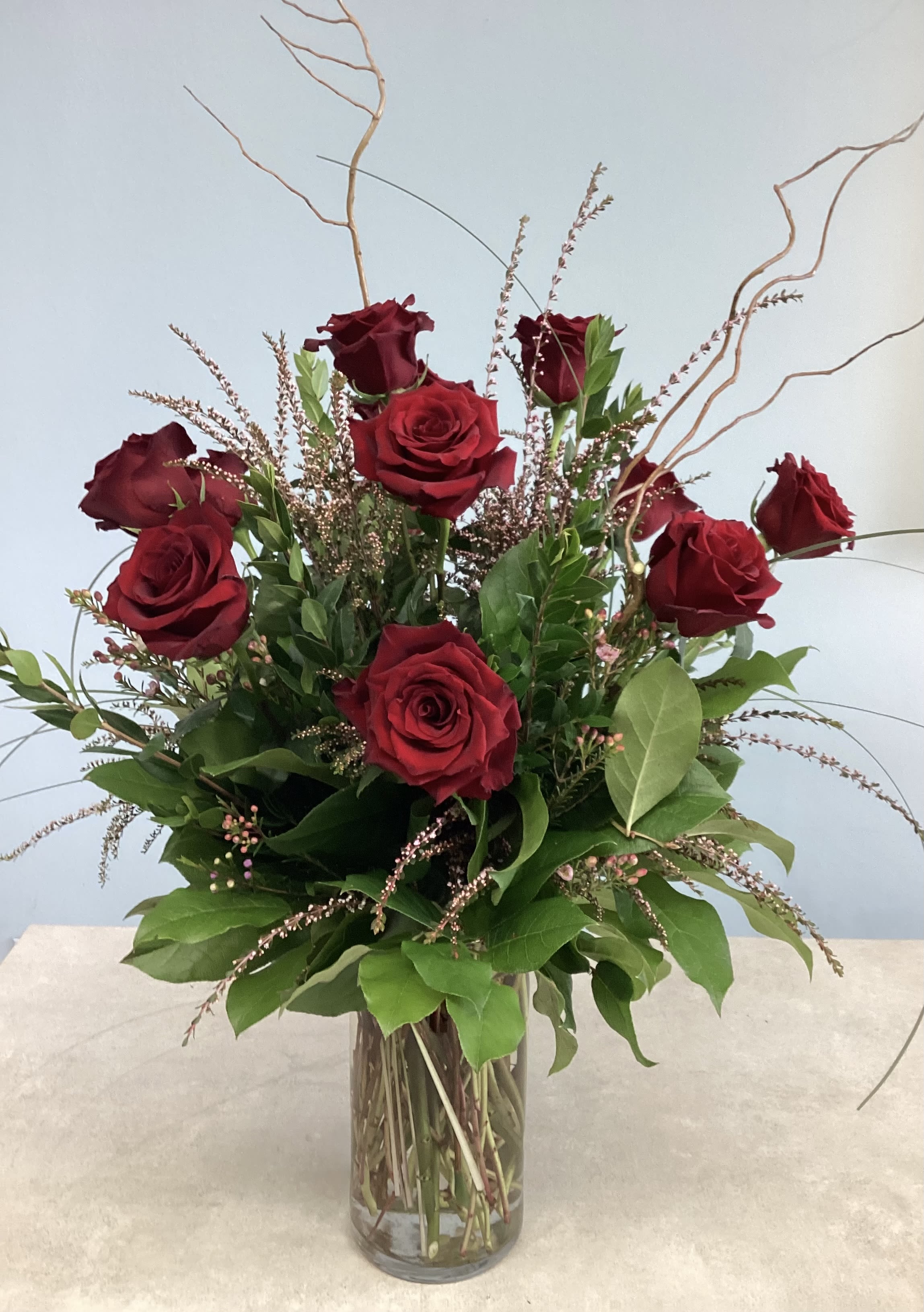 Dozen Red Roses - Premium, deep red roses with curly willow, lush green foliage and filler presented in a clear glass vase - a romantic, classic arrangement perfect for anniversaries or thoughtful everyday gifting.