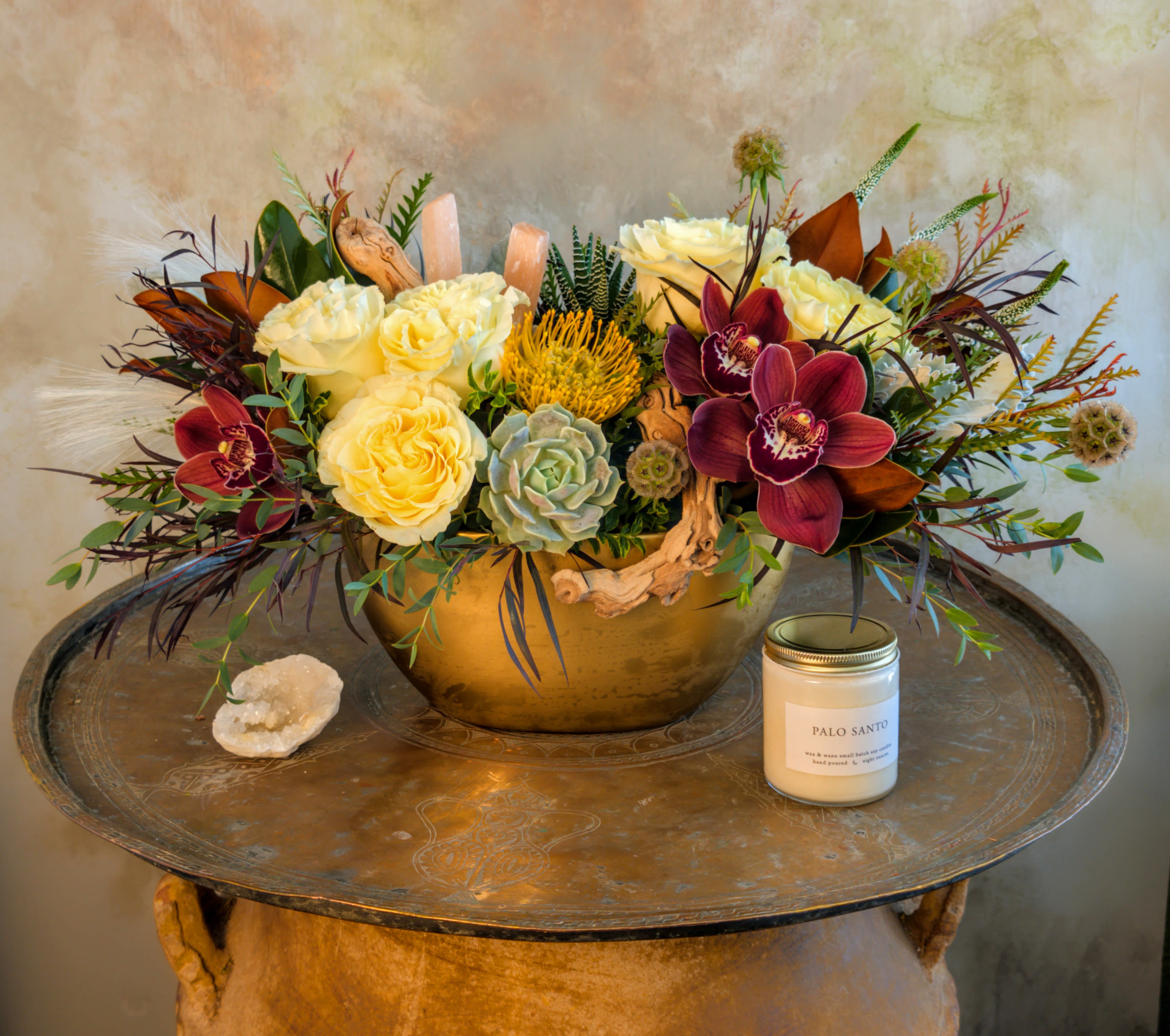 Arrangement of cream roses and burgundy orchids in a gold bowl with a candle