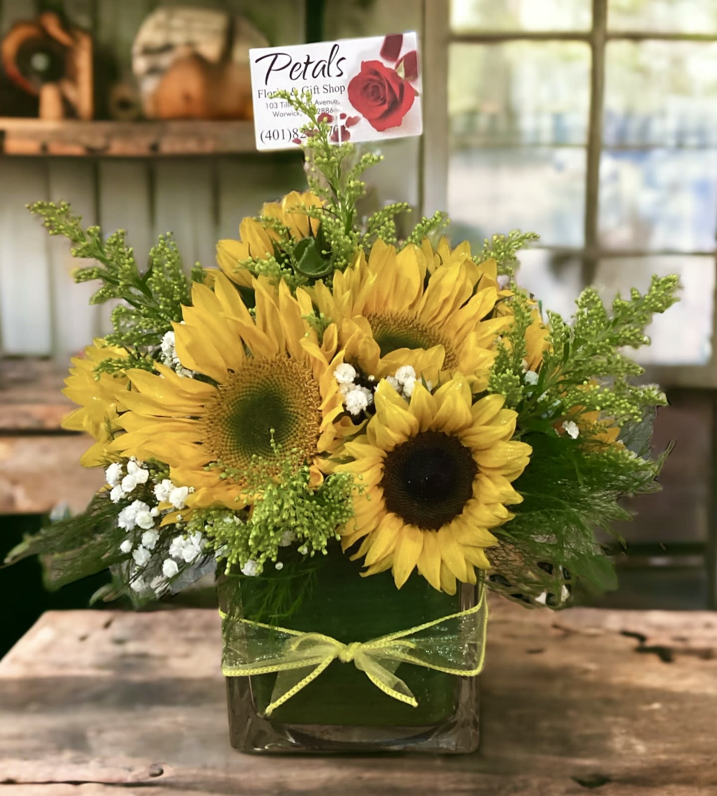 Sunflowers &amp; Solidago Cube by Petals - Capture a moment of pure bliss with the Sunflower &amp; Solidago Cube. This charming floral display is a story in bloom, with sun-kissed sunflowers and their golden companions gathering together to bring a piece of the summer garden indoors. The 5x5" clear glass cube acts as a frame, allowing you to witness a daily dose of happiness. Give the gift of a smile, and watch as this bouquet becomes a beloved centerpiece of joy.    