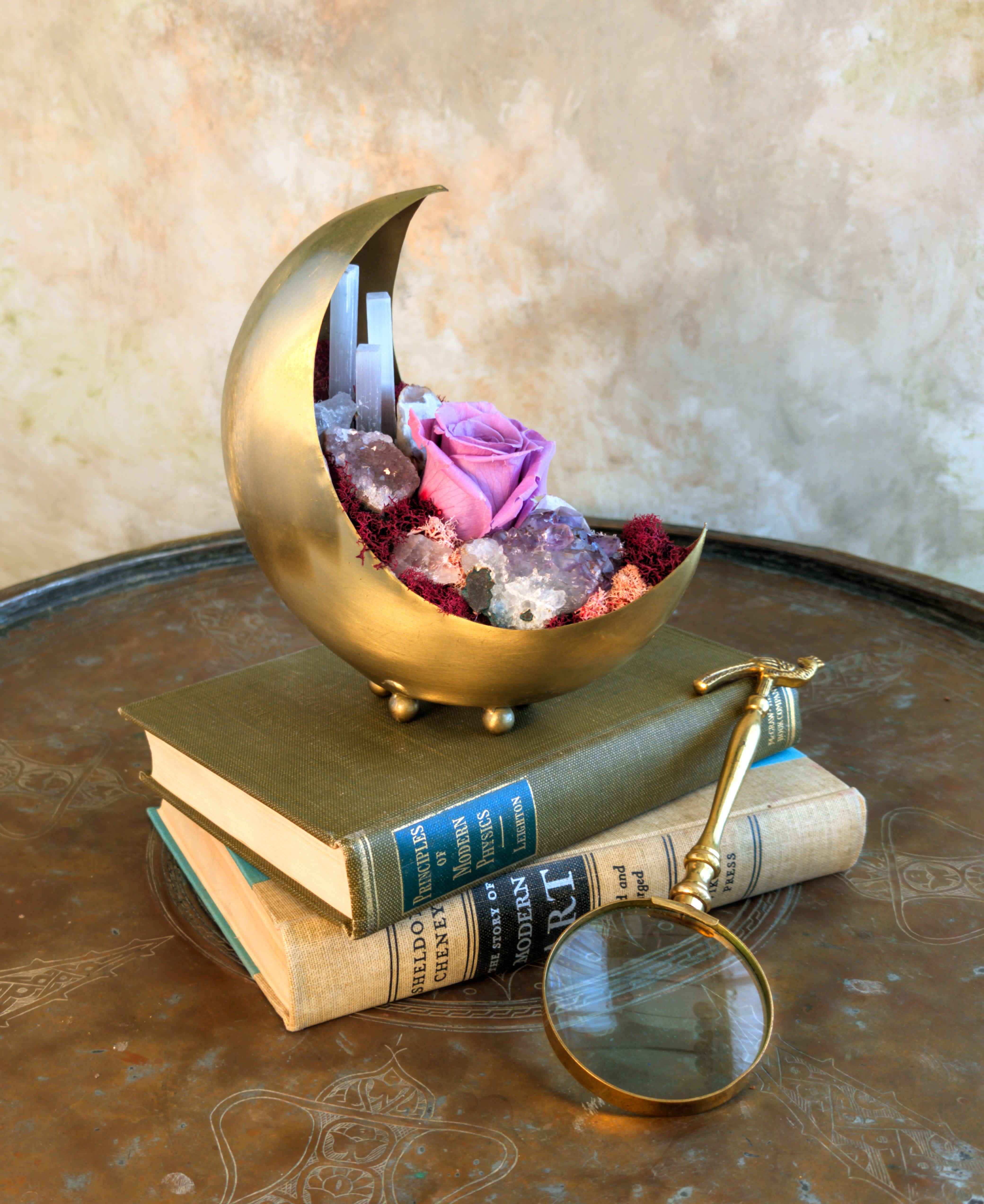 Pink rose in a gold crescent-shaped container with crystals and books
