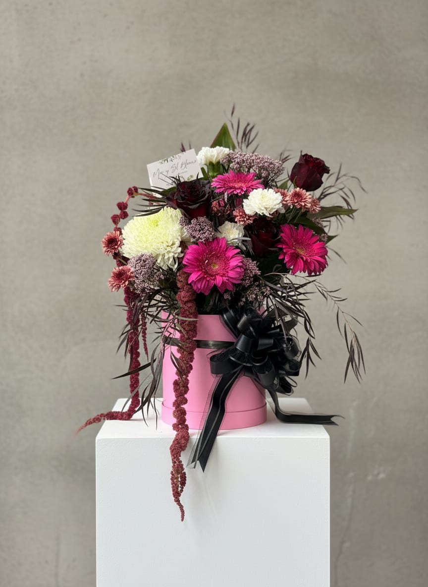 Moody Pink - A bold and romantic arrangement designed to make a statement. Featuring rich burgundy roses, vibrant fuchsia gerbera daisies, delicate blush blooms, and crisp white chrysanthemums, this design is artfully arranged in a chic pink box and finished with an elegant black ribbon.  Our hanging amaranthus and textured foliage add movement and depth, creating a modern, luxurious look with a touch of whimsy. Perfect for anniversaries, birthdays, celebrations of love, or simply reminding someone how special they are.