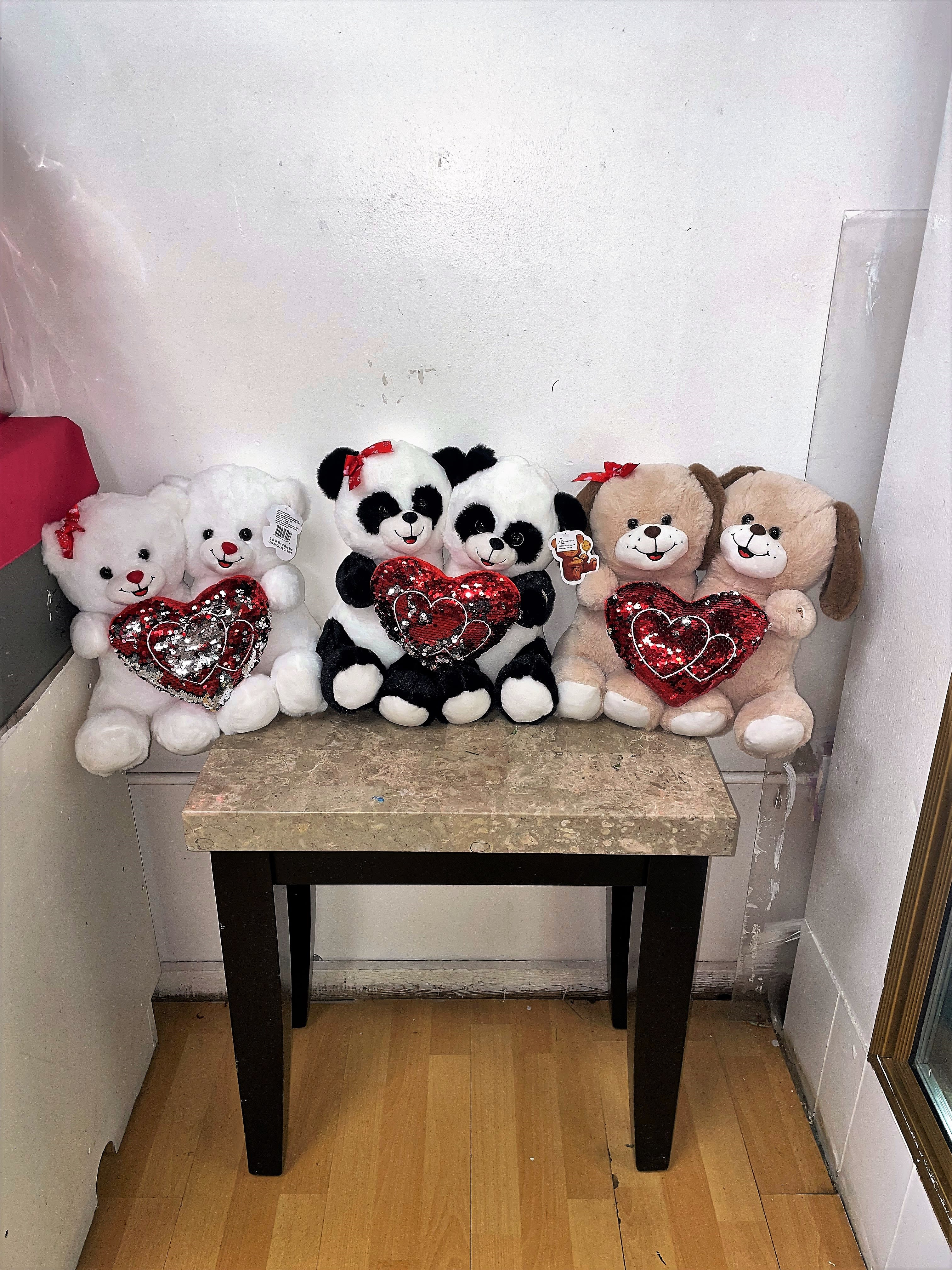 Three plush teddy bears and two panda plush toys holding red heart pillows on a table.