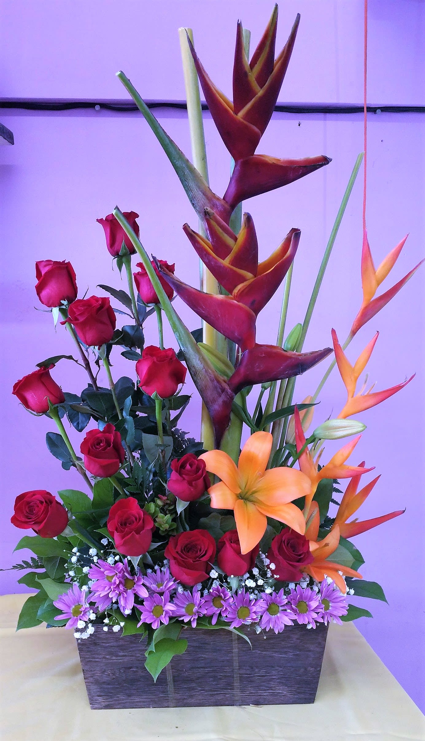Tall mixed flower arrangement with red roses, orange heliconia, and purple daisies in a rectangular container