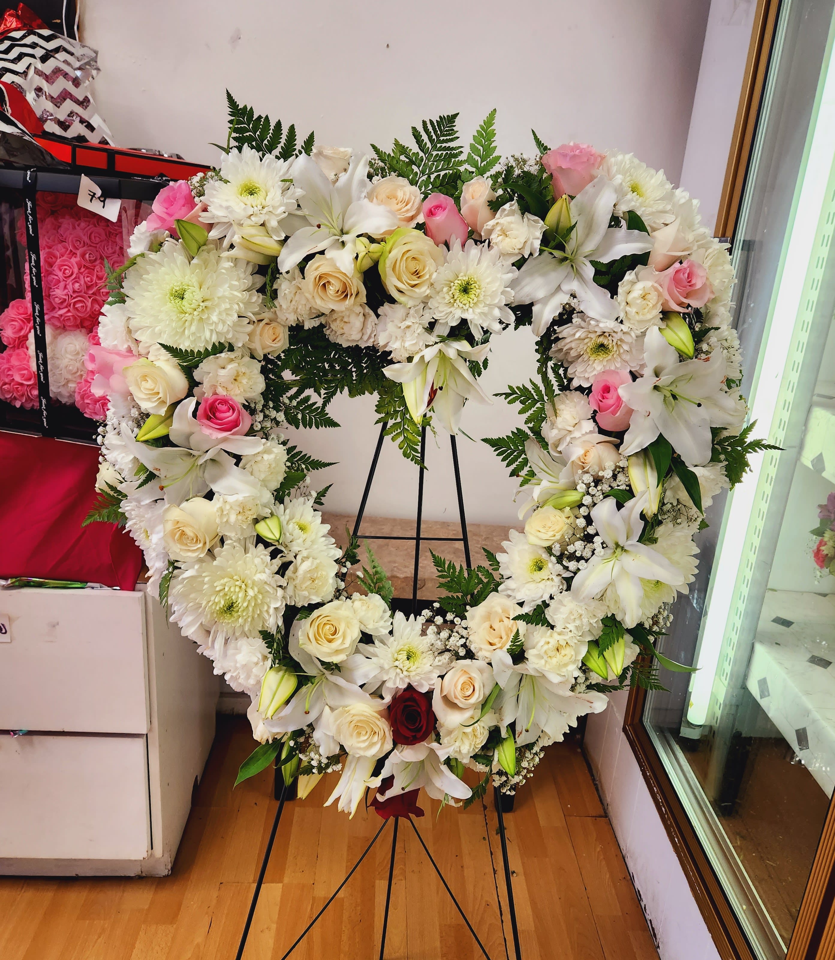 Heart-shaped floral wreath with white lilies, roses, and pink accents on a stand