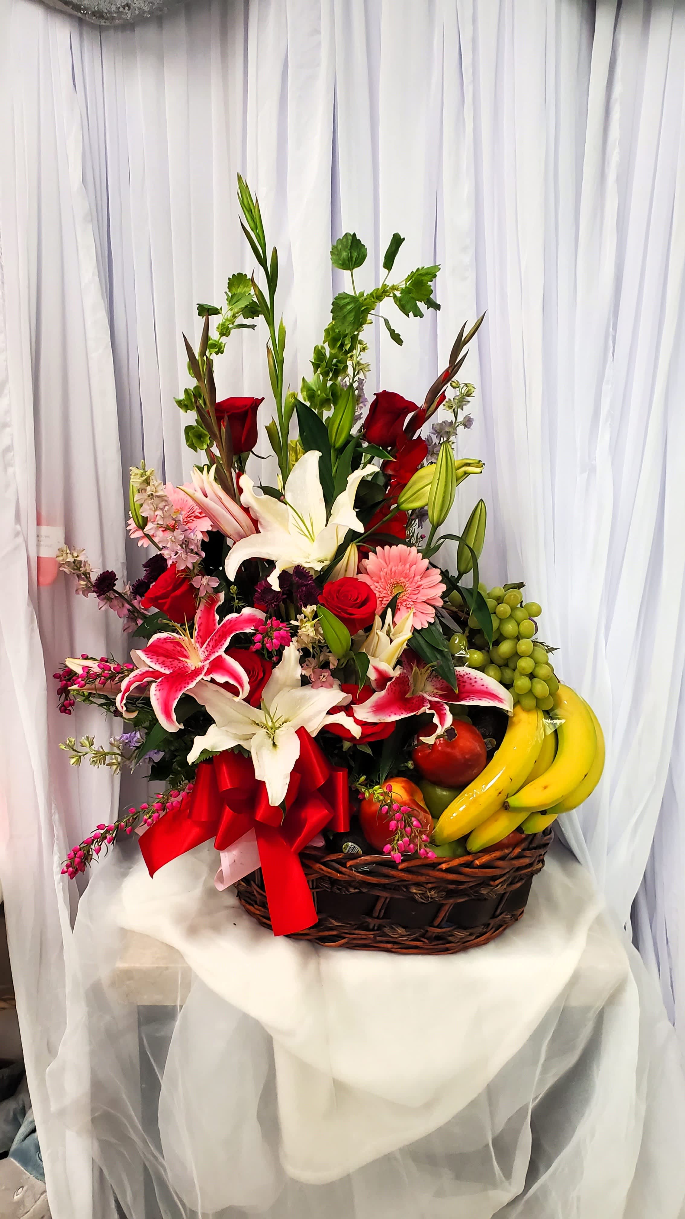 Basket arrangement of lilies, roses, and fruit with a red ribbon