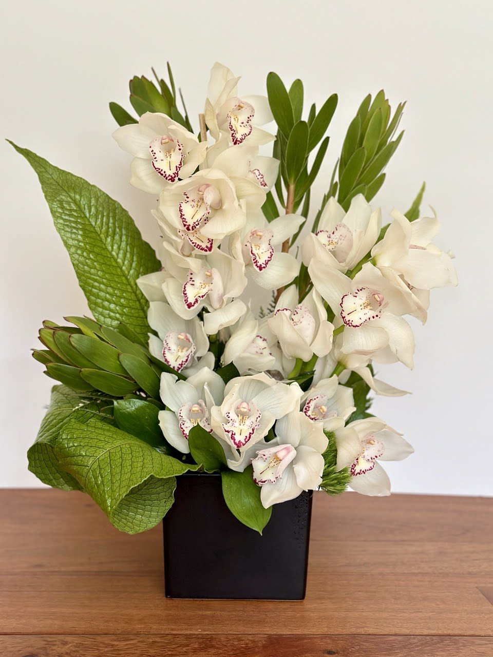 Arrangement of white orchids with burgundy centers and large green leaves in a black cube vase