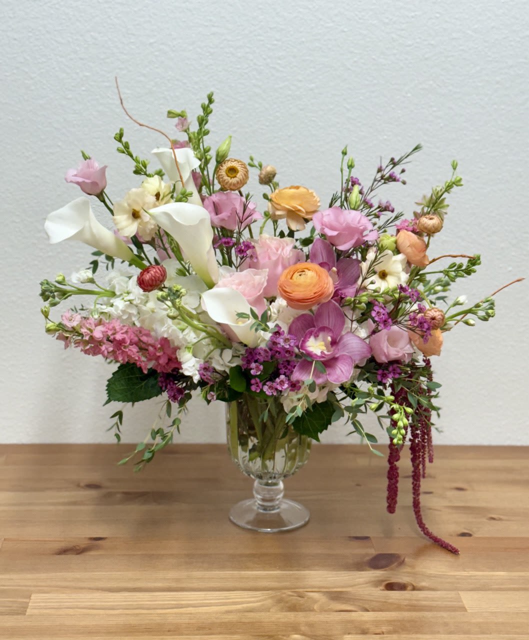 Asymmetrical pastel arrangement with calla lilies, ranunculus, orchids, and mixed blooms in a clear glass vase
