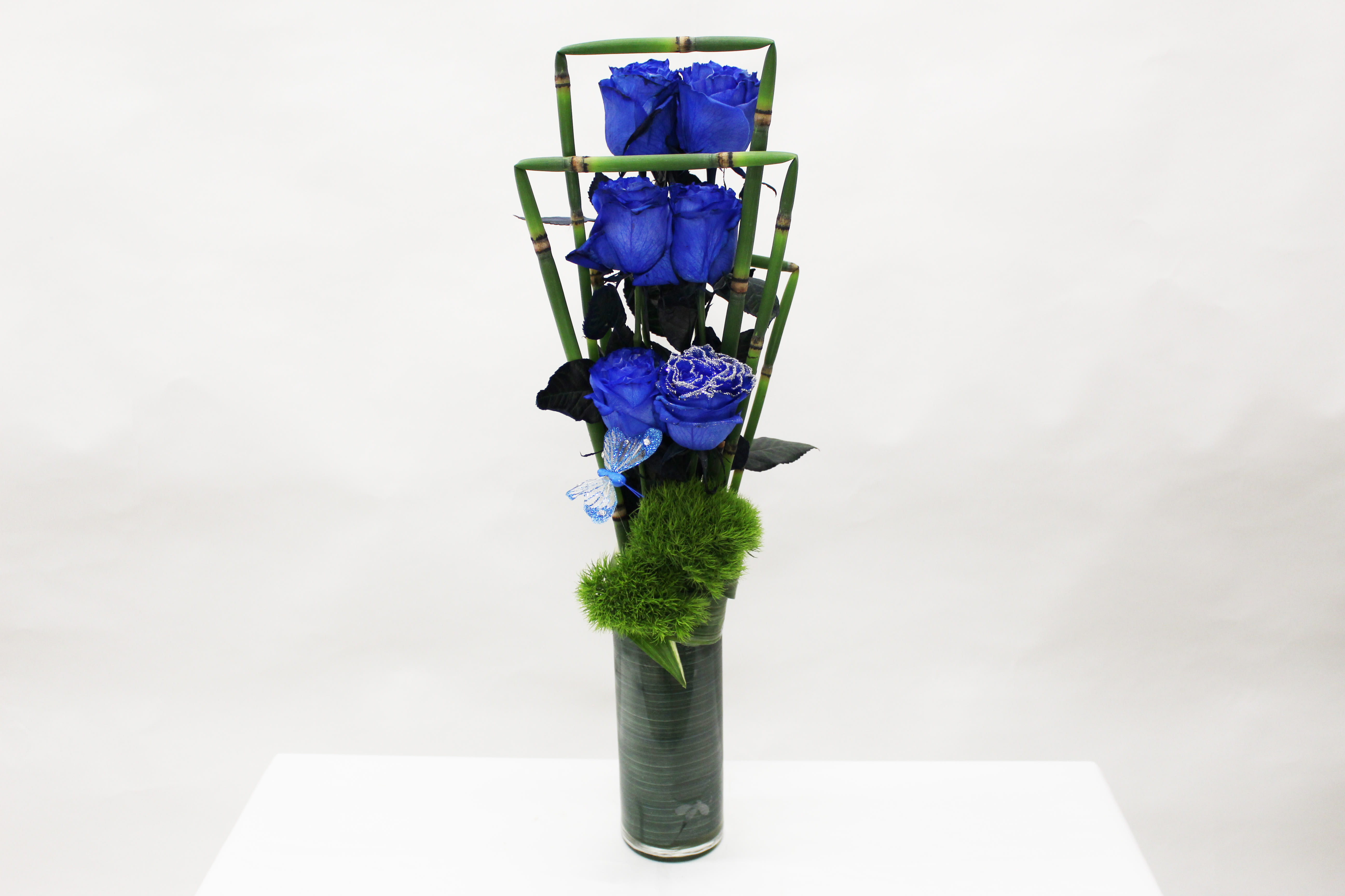 Tall modern arrangement of vivid blue roses in a green-lined glass cylinder with bamboo accents and fuzzy green dianthus