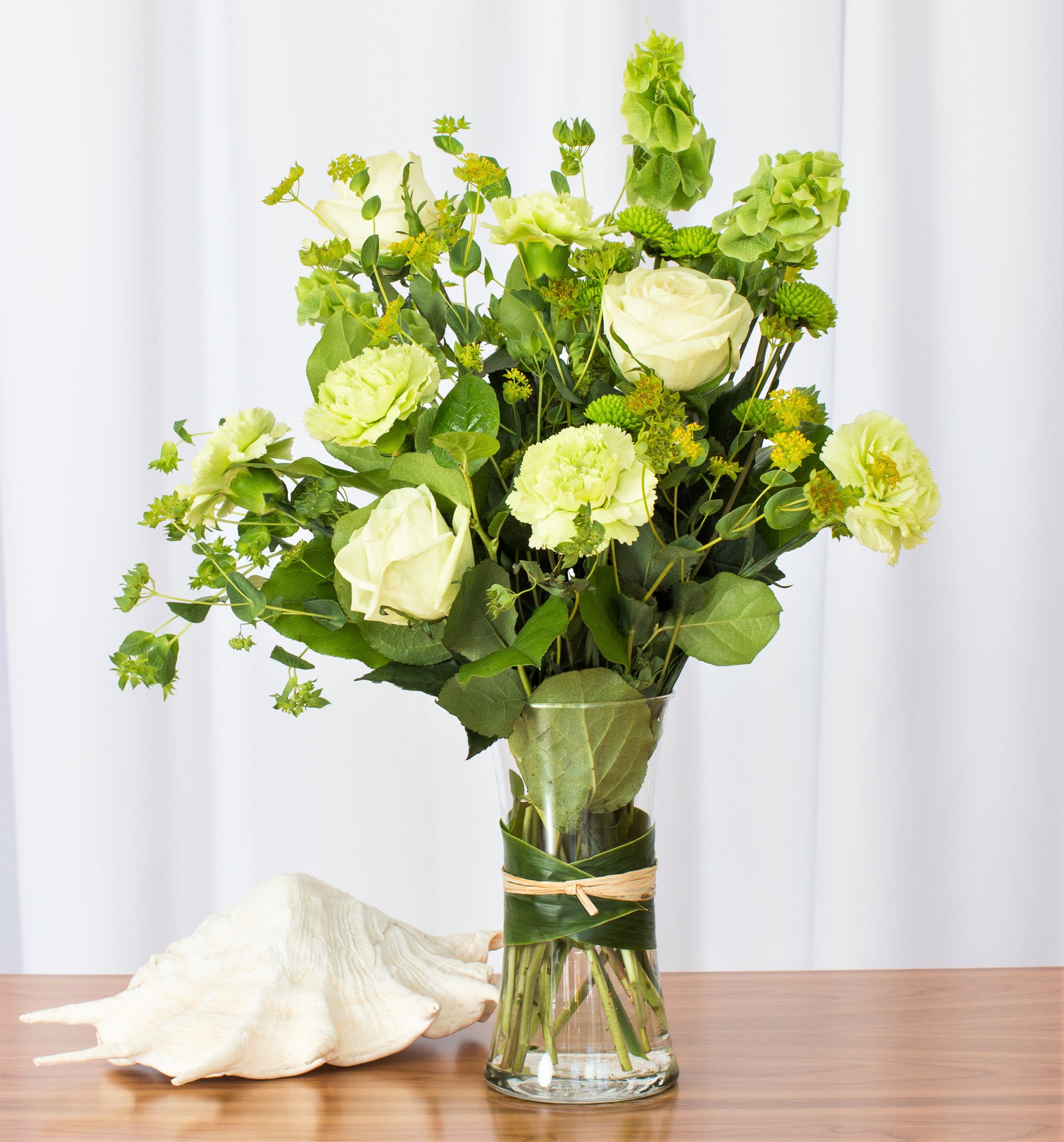 Tall arrangement of pale green roses and carnations in a clear vase wrapped with leaves beside a seashell.