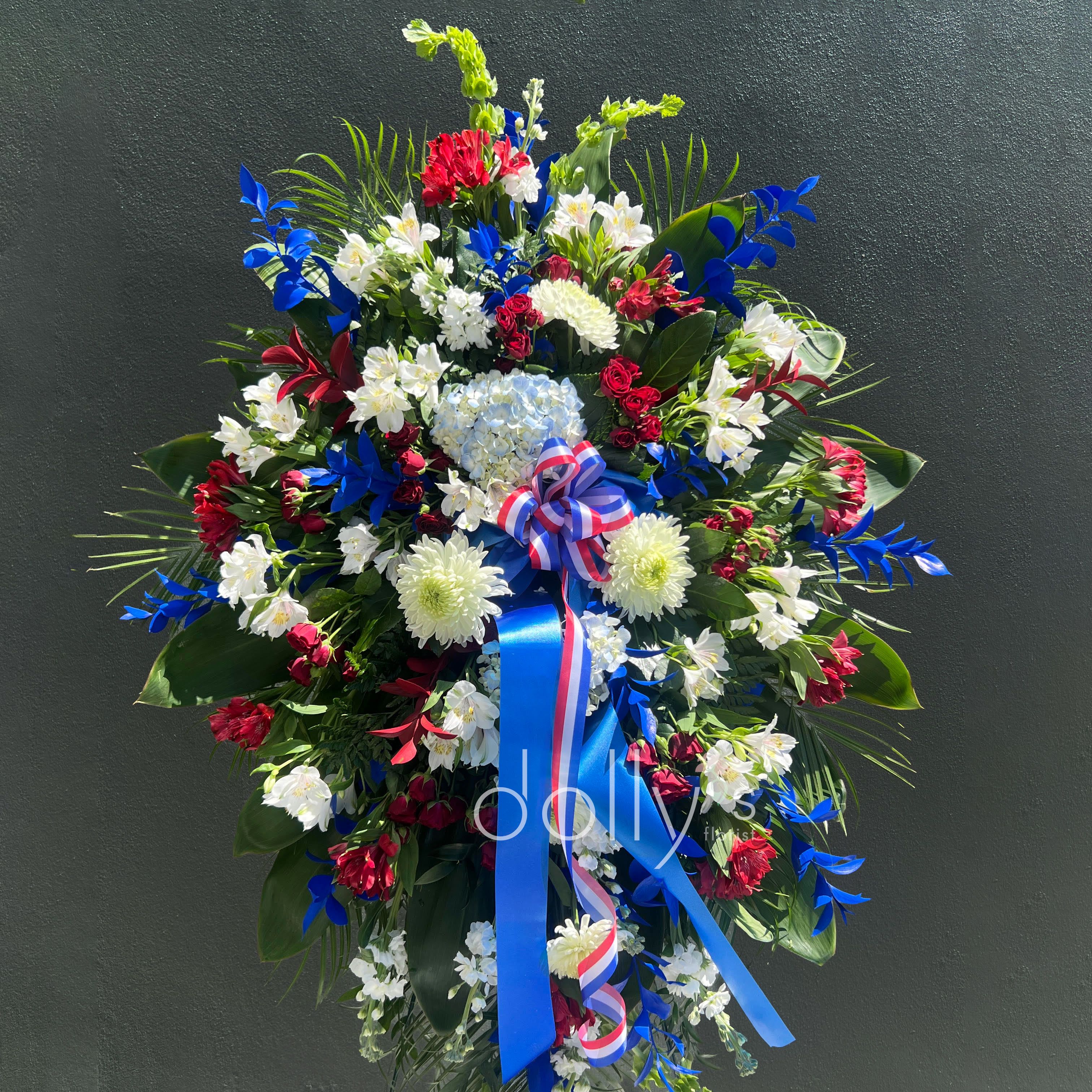 Our Patriot - A commanding standing sympathy spray that honors a life with both patriotic pride and tender remembrance - perfect for a funeral, memorial service, or commemorative ceremony. The arrangement reads as a formal memorial tribute that will help the person you're remembering feel respected and seen.  This large easel spray is built with a dense, layered mix of chrysanthemums, roses, lilies, carnations, alstroemeria, spray roses, hydrangea and daisy-like fillers set over broad palm fronds and glossy greenery. The red-white-and-blue style highlights clusters of blue hydrangea with white chrysanthemums and red accents and is finished with blue and red-white-and-blue ribbon streamers. The piece is tall and substantial with a rounded, slightly cascading silhouette and spiky outer texture that reads well from a distance in a chapel or reception space.  Choose this spray when you need a dignified, expressive tribute that matches a patriotic service or a gentle memorial tone. It arrives on a standing easel or frame ready for display; include your sympathy card message at checkout and our designers will attach the selected ribbon and card for a finished presentation appropriate to the service.