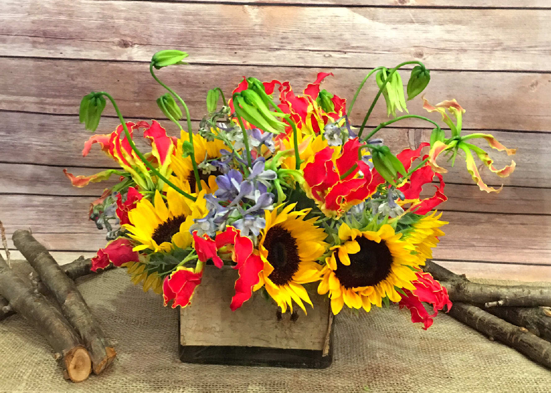 BLAZING BEAUTY  - EXTRAVAGANT BLEND OF GLORIOSA LILY, SUNFLOWERS, AND BLUE DELPHINIUM ARRANGED IN SQUARE GLASS LINNEN WITH WHITE BIRCH BARK. 