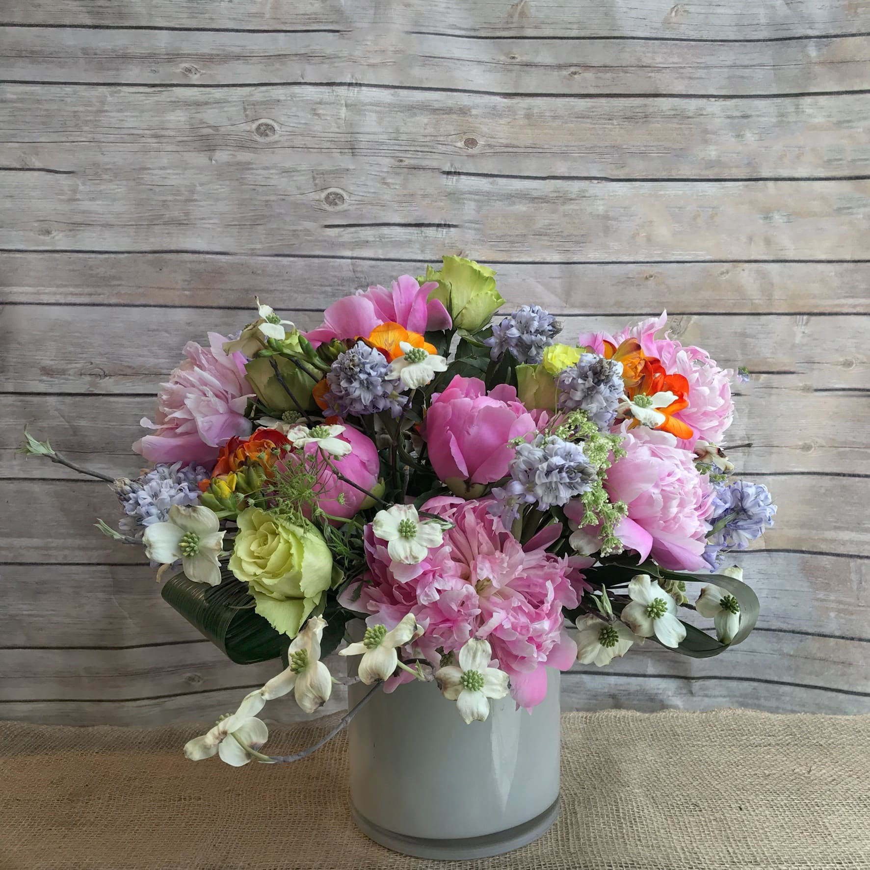 EVERYTHING I AM  - ASSORTMENT OF SILA HYACINTH, WHITE DOGWOOD, ORANGE FREESIA, GREEN ROSES,AND JOULES ELSIE PEONIES IN A MODERN WHITE GLASS CYLINDER VASE.