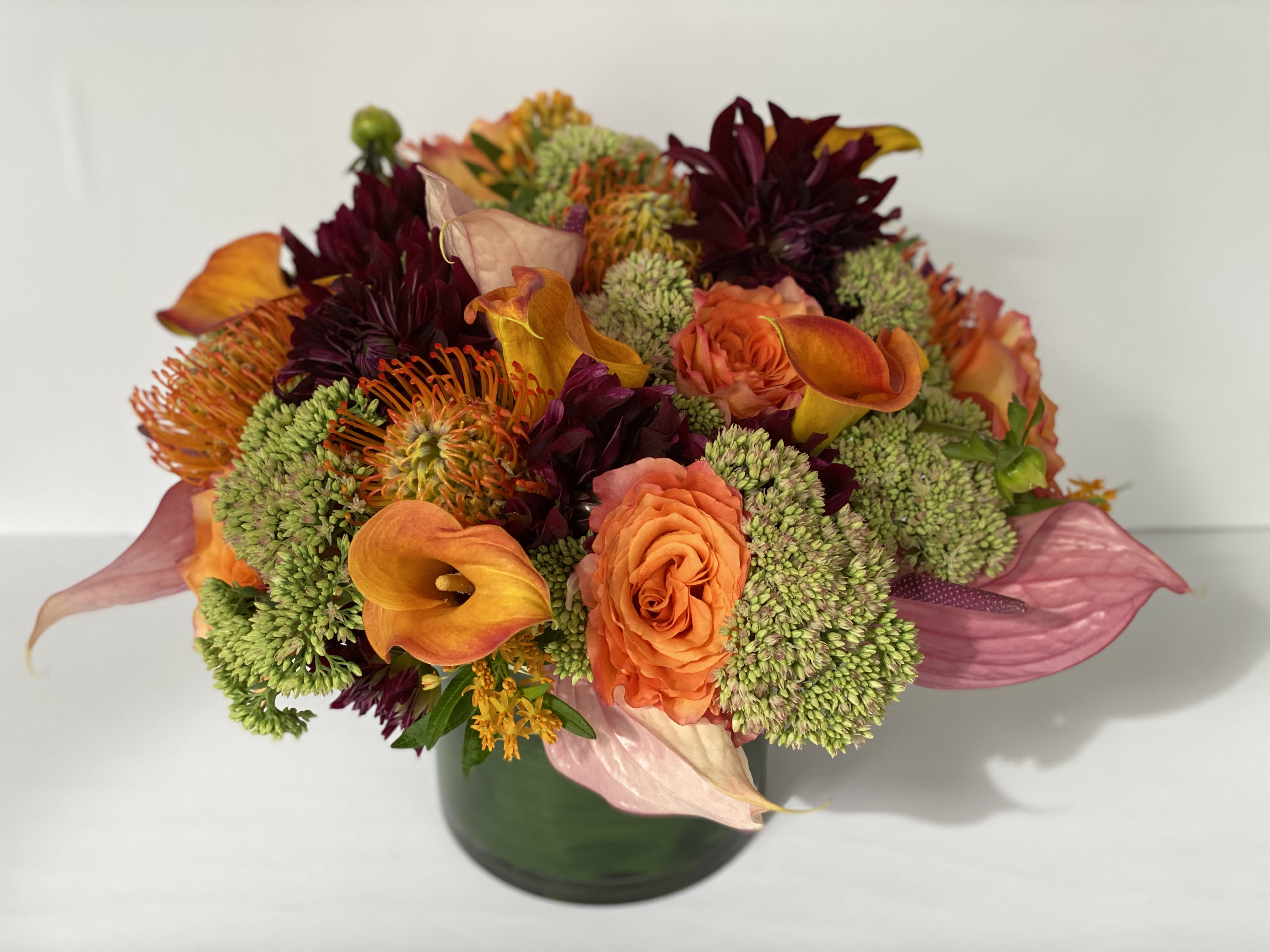 LUXE JEWEL - LOW AND FULL ROUND BOWL WITH A SELECTION OF FREE SPIRIT ROSES, DAHLIAS , CALLA LILY'S, SEDUM, PIN PRO TEAS AND ANTHURIUMS FLOWERS IN AUTUMN TONES.  