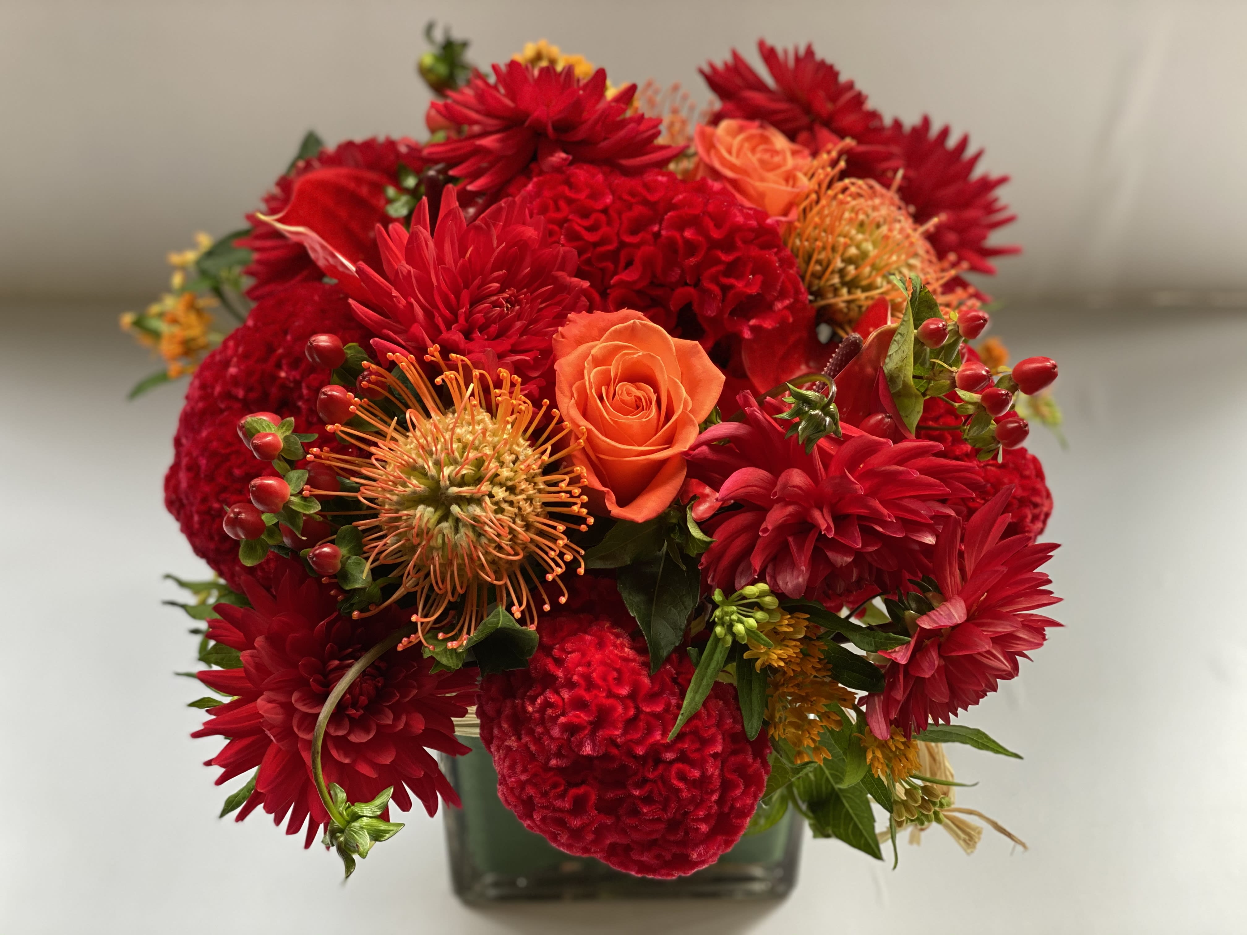 FIRE ISLAND  - This lush composition is surrounded of  dhalias karma bon bini, crush orange roses  , nutan pro-tea, asclepias in oranges, velvety celosia,  in a low cube or round cylinder. 