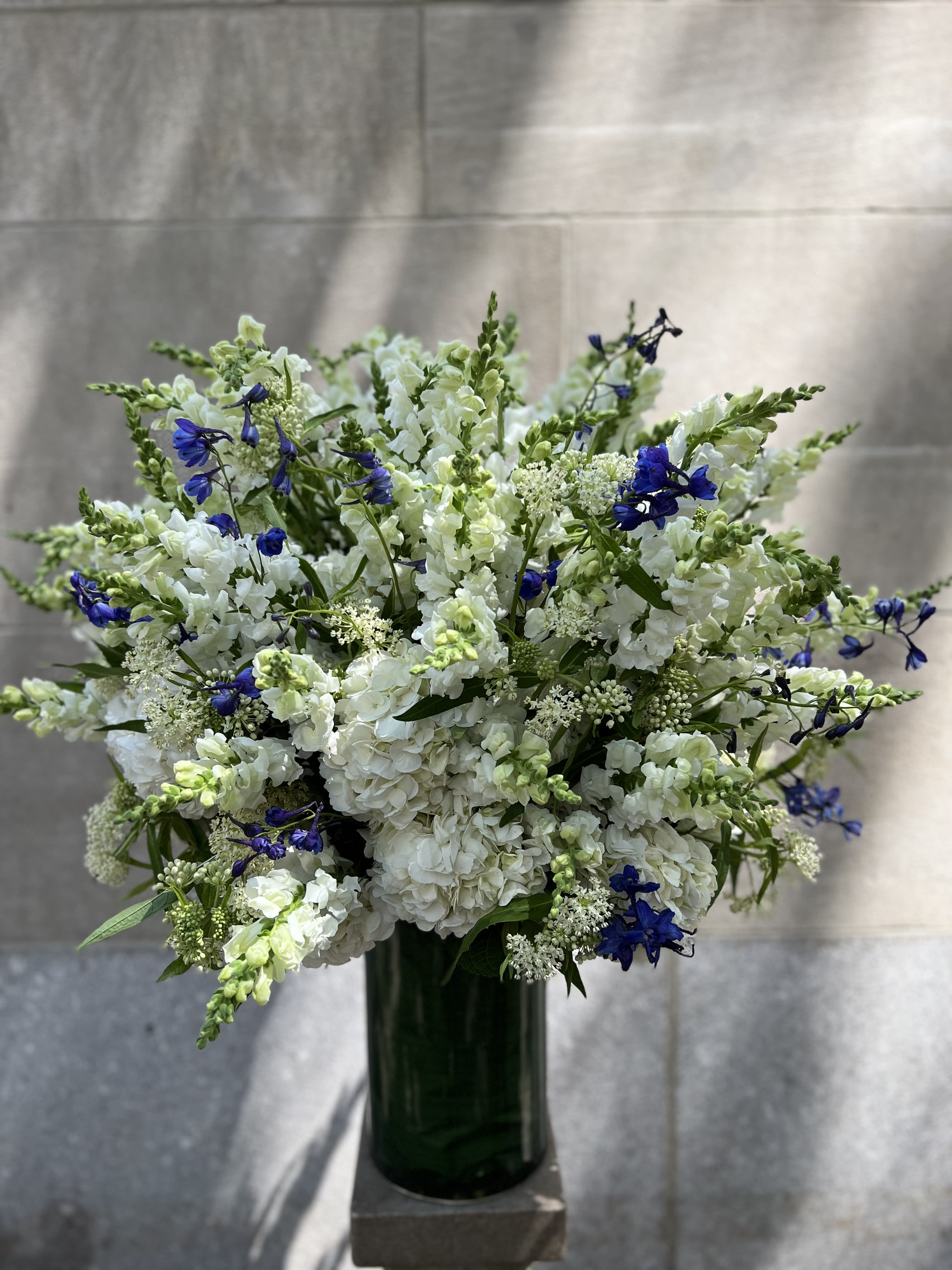 BEMA - For the service: Features a lush assortment of primarily white color flowers that includes: hydrangeas, asclepias, snap dragons tucked in with a rich blue color delphinium flower. Display measures: 30"H * 24" W 