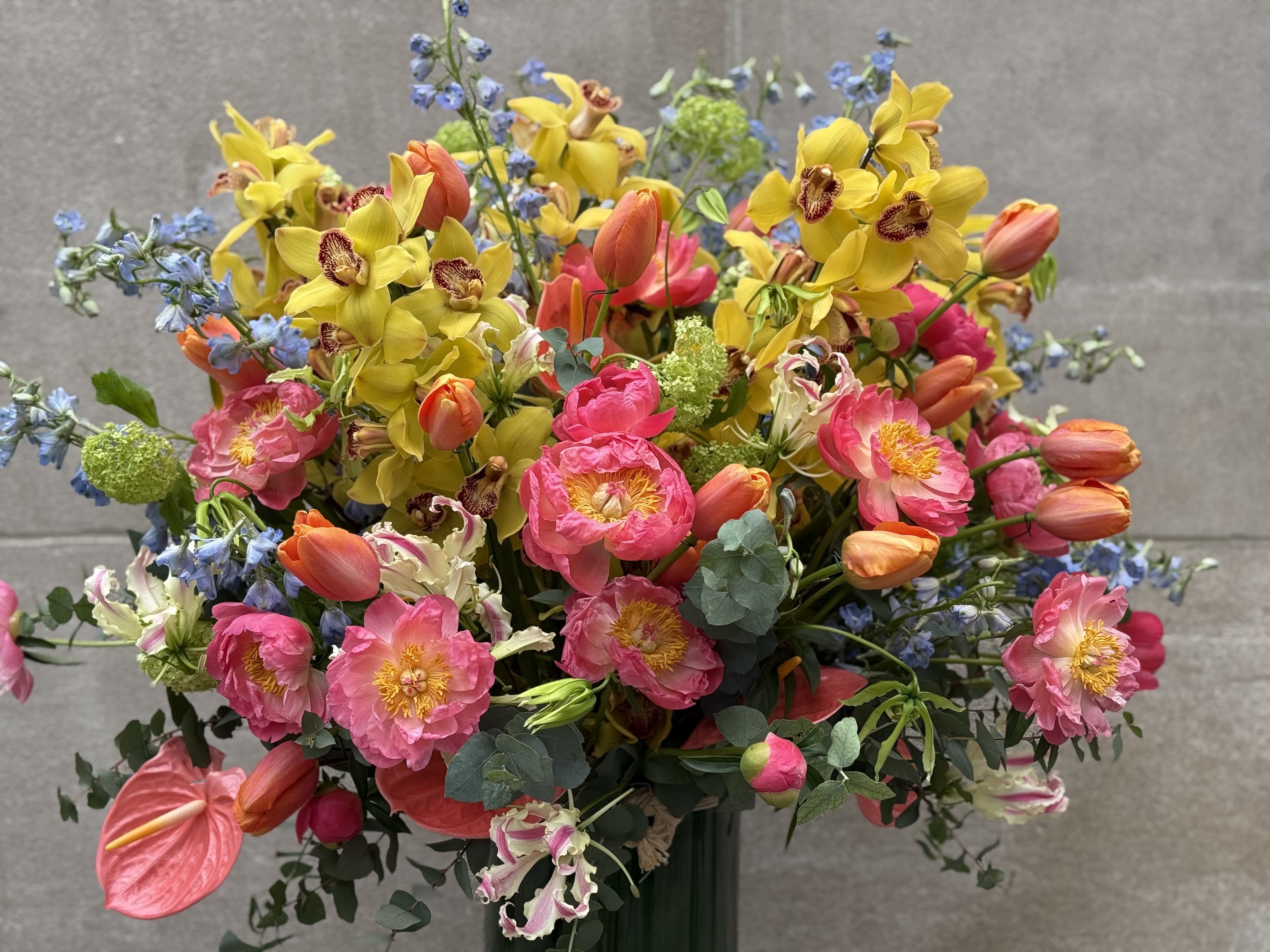 Tribeca - Flowers by Philp proudly presents this stunning and abundant composition featuring coral peonies, cheerful yellow cymbidiums, and elegant delphiniums! Get ready to be dazzled by this large, vibrant, and lush assortment that will brighten any space!
