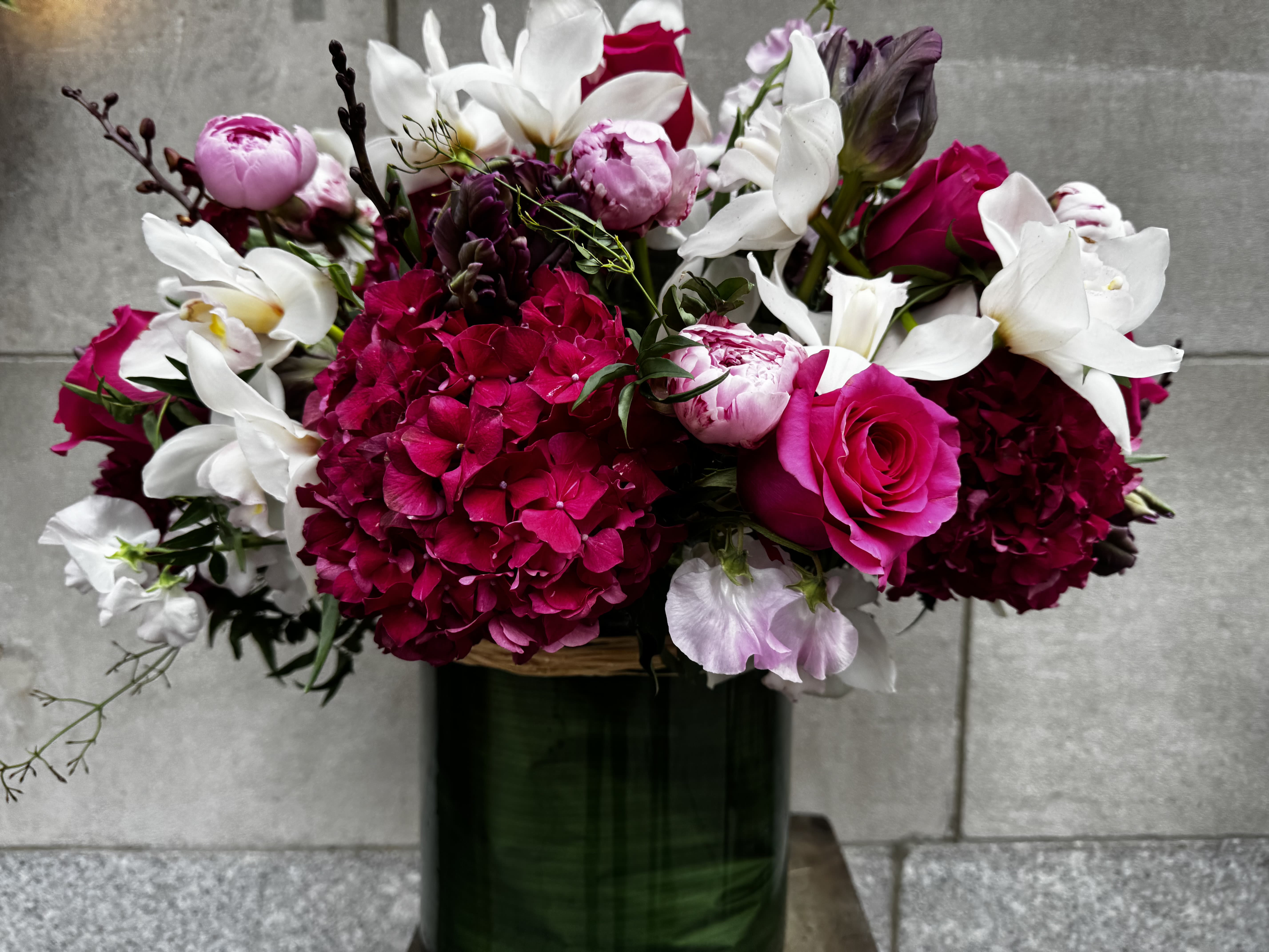 LOLA - Get ready to have your senses tickled by this floral masterpiece that looks like it just walked off a fancy runway! We’ve got Colombian hydrangeas strutting their stuff, white cymbidium orchids that could make a dessert jealous, pink Floyd roses ready to rock your world, sweet peas that are sweeter than your grandma's cookies, and parrot tulips that are just a little too colorful for their own good—all hanging out together in a low round cylinder. It’s like a botanical party that even your houseplants would be jealous of!