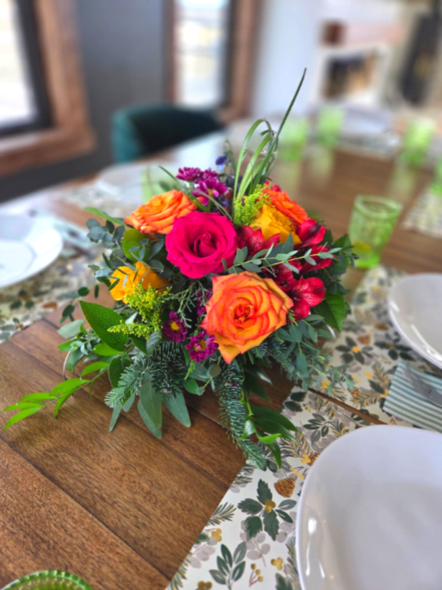 Spring has Sprung Centerpiece  - A vivid dome of orange, pink, yellow and purple petals greets the room-this low floral arrangement is a spring centerpiece made for birthdays, congratulations, or simply letting someone know you're thinking of them. The rounded, slightly spilling silhouette sits close to the table so the person you're celebrating can easily enjoy the saturated colors and joyful energy across a dining or coffee table.  Clusters of bi-color orange-and-yellow roses are paired with hot pink roses, purple and deep magenta accent blooms, alstroemeria and chrysanthemums woven with small yellow filler flowers and layered greenery. The compact design is arranged in a low floral container that keeps the piece intimate and conversation-friendly while giving a dense, textured look that reads lively from every angle.  Perfect for festive gatherings or everyday appreciation, this centerpiece makes the recipient feel celebrated with bright, uplifting color. Sits low for easy placement on tabletops; keep water fresh and trim stems as needed to prolong freshness.