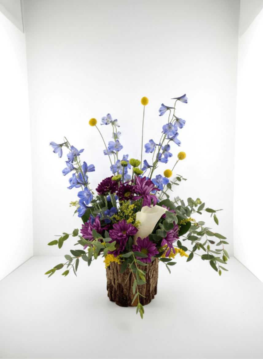 Fresh Forest - Tall blue delphinium-like spikes paired with purple daisy-style blooms make this Fresh Forest flower arrangement feel like a sunlit corner of the garden - perfect as a birthday bouquet, a cheerful everyday gift, or a sincere thank-you arrangement. The airy, whimsical silhouette lets the taller stems rise above a compact base so the recipient will notice movement and height the moment they enter the room.  Soft white chrysanthemums and small yellow button flowers mix with eucalyptus-like foliage for contrast in both color and texture, while the daisies keep the mood light and friendly. A rustic wood container grounds the design and is offset by a white floral wrap tucked into the center, giving the arrangement warmth and a handcrafted touch. Dimensions and exact flower counts vary by size selection; the composition is designed to spill outward naturally and arrive ready to display.  This is the kind of arrangement that makes someone feel genuinely seen - bright and uplifting without being formal. For best longevity, keep in fresh water away from direct heat and replace water every other day; choose your preferred size at checkout to match the occasion and the recipient's space.