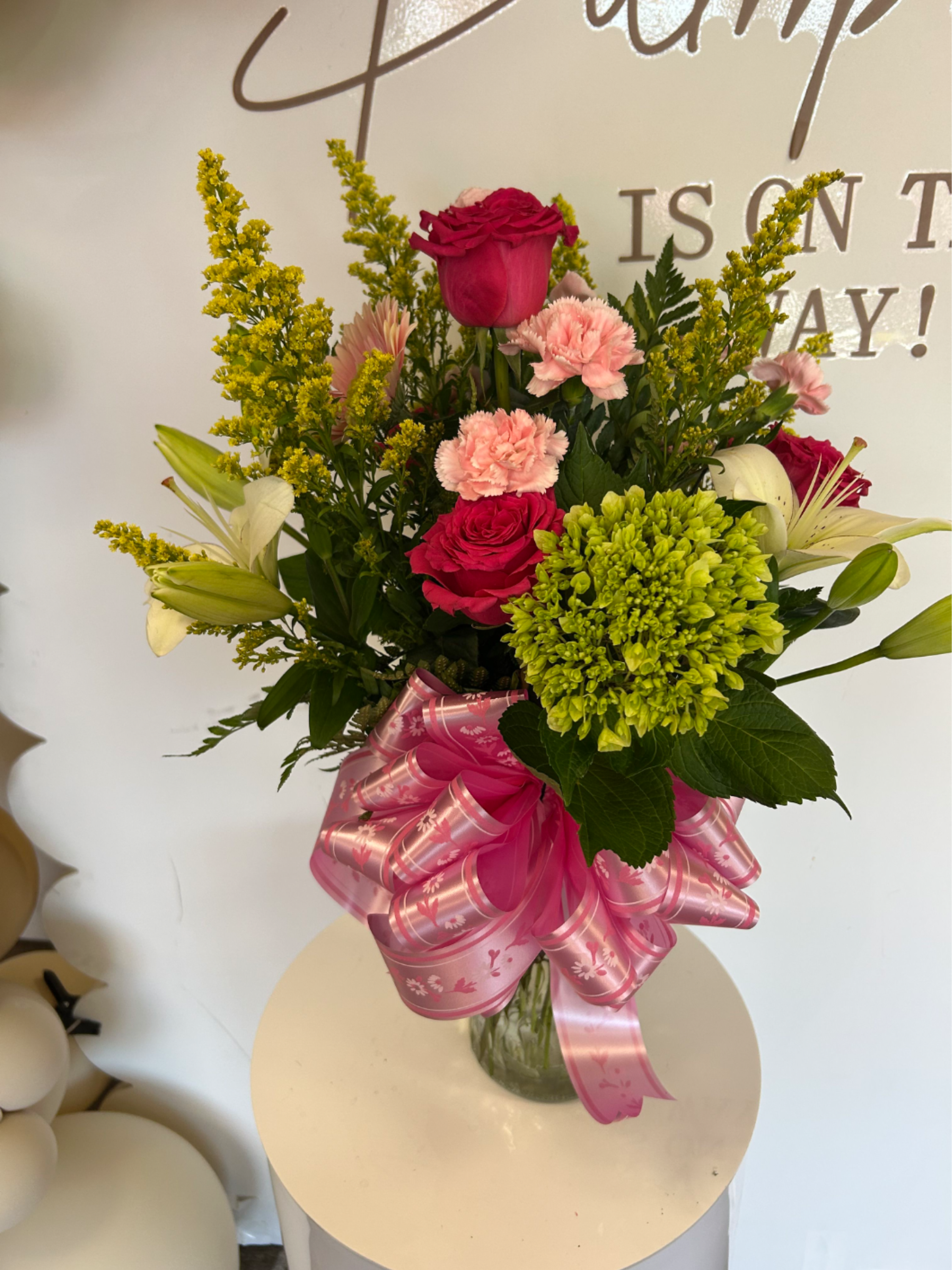 Pink glam  - Bright, celebratory pink roses and pale pink carnations take center stage in this flower arrangement, a perfect birthday bouquet or thoughtful thank-you gift that feels as festive as it looks. White lilies and pops of yellow filler brighten the composition while a lush green hydrangea cluster (in some variations) adds volume and a fresh contrast-so the person you're sending it to will feel genuinely celebrated and seen.  Arranged in a clear glass vase and finished with a broad pink decorative ribbon bow at the base, this design balances saturated pinks with crisp whites, sunny yellow accents, and green texture for a lively, romantic undertone. The mix of roses, carnations, lilies, hydrangea, and delicate filler flowers gives the bouquet a full, multi-layered look and a pleasing range of shapes and scents.  Ideal for birthdays, congratulations, or a feminine everyday surprise, this gift-ready arrangement arrives styled and tied with care; simply place in a cool spot away from direct sunlight and refresh the vase water every 2-3 days to keep blooms looking their best.
