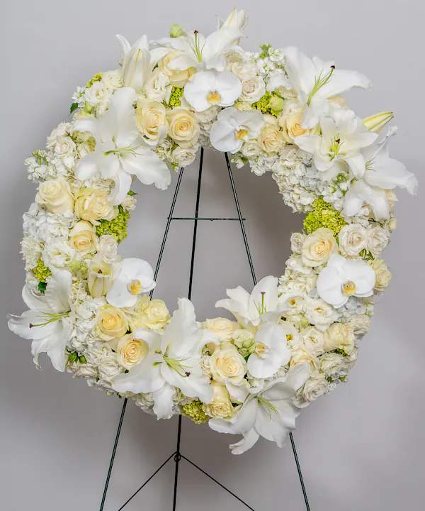 Graceful In White wreath - Their pure heart and peaceful spirit will live on in a gesture symbolizing the circle of eternal life. Our standing wreath of soothing white blooms and fresh greenery, expertly crafted by our local florists for a lush, full presentation, is a grand expression of your condolences, offering hope and serenity during a difficult time.. elegant wreath created from premium white blooms including hydrangea, lilies, orchids, and roses.
