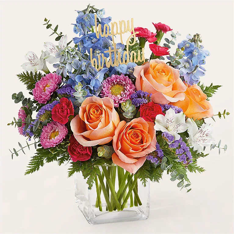 Mixed bouquet of roses, daisies, and blue flowers in a clear vase with a birthday sign
