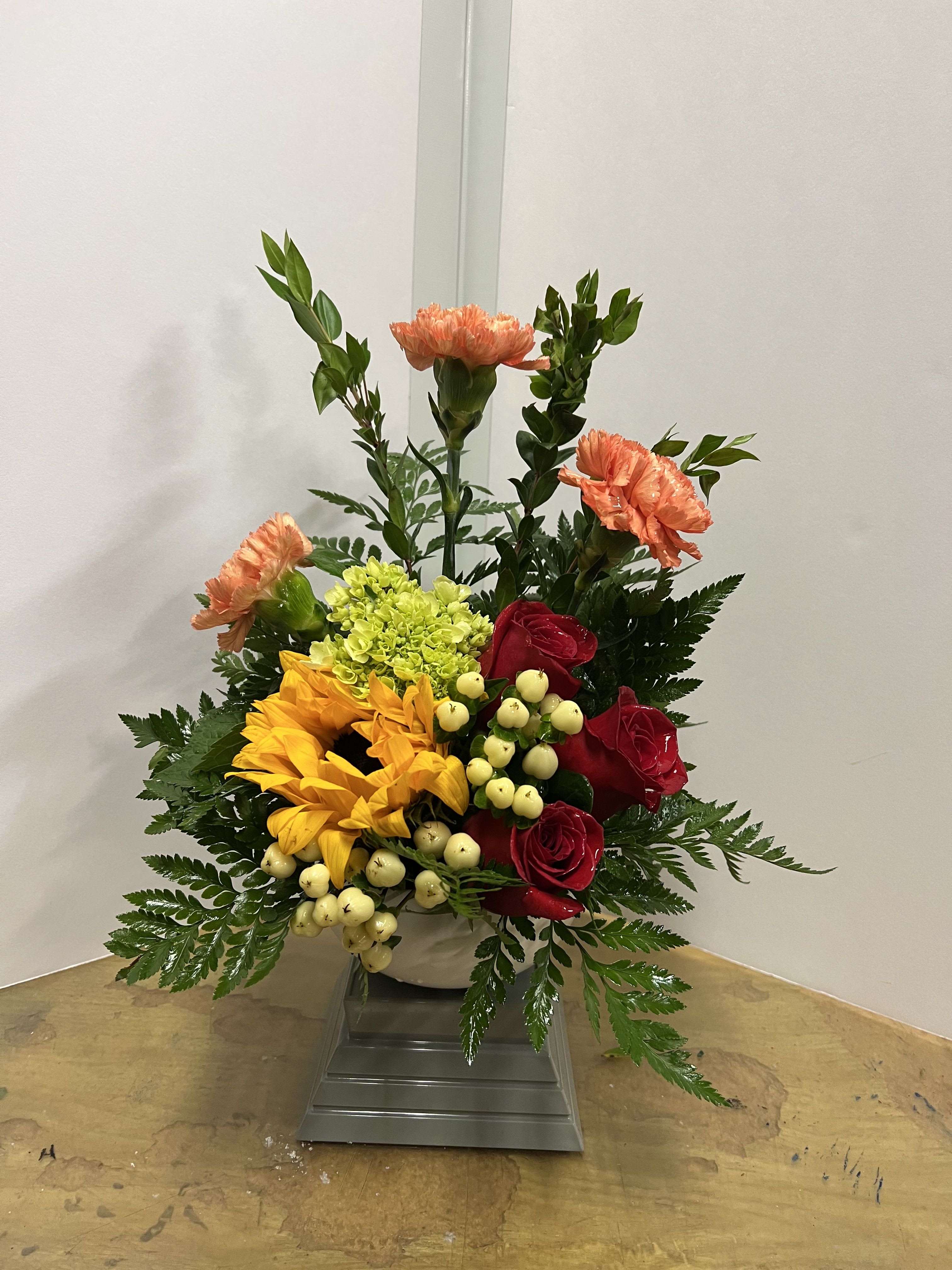 Mixed bouquet with red roses, yellow sunflowers, and peach carnations in a white vase