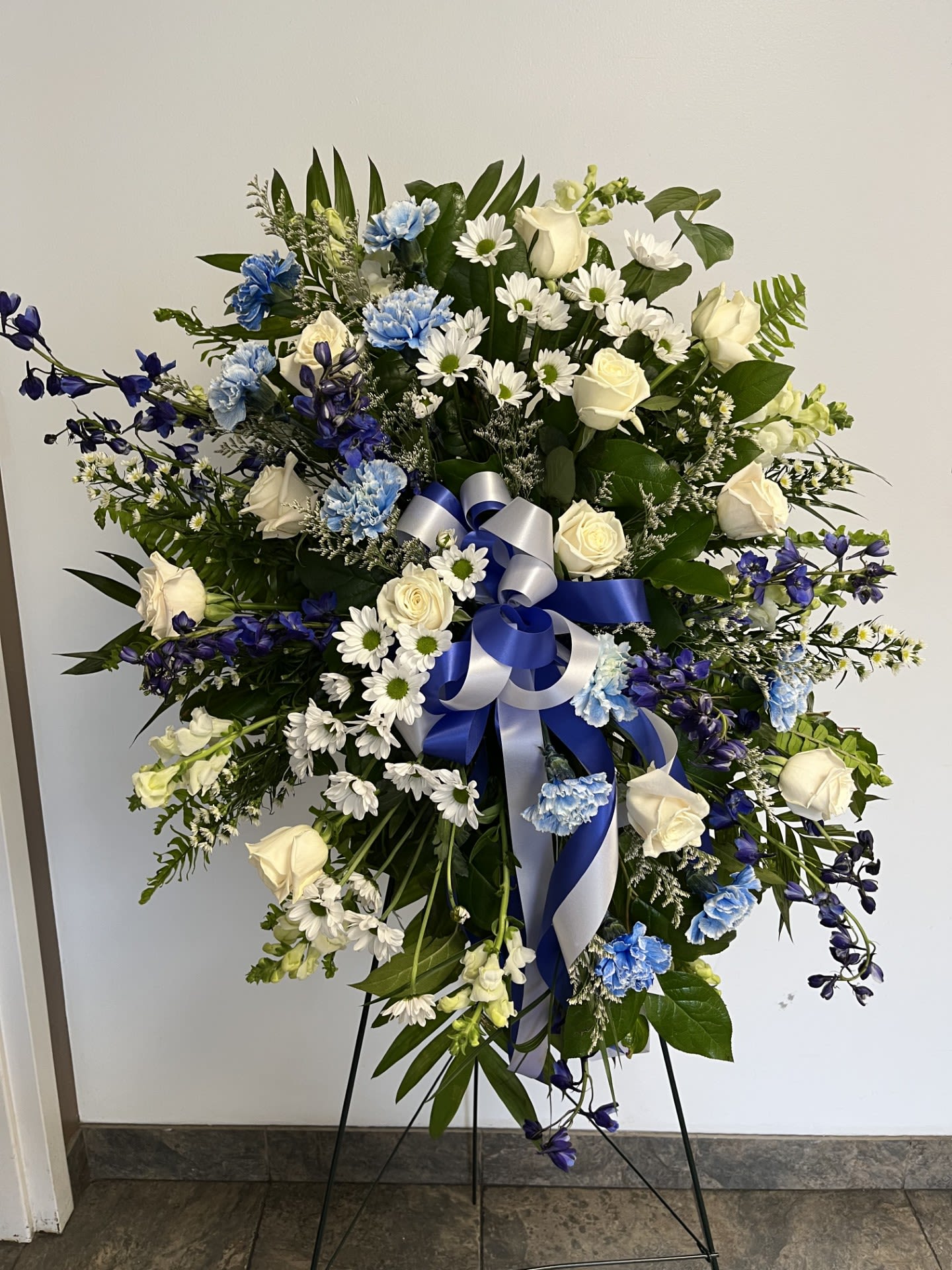 Tall blue and white standing spray with roses, carnations, daisies, and a blue and white bow on an easel.