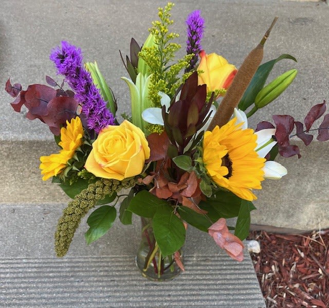 Mixed bouquet with yellow roses, sunflowers, and purple spikes in a glass vase