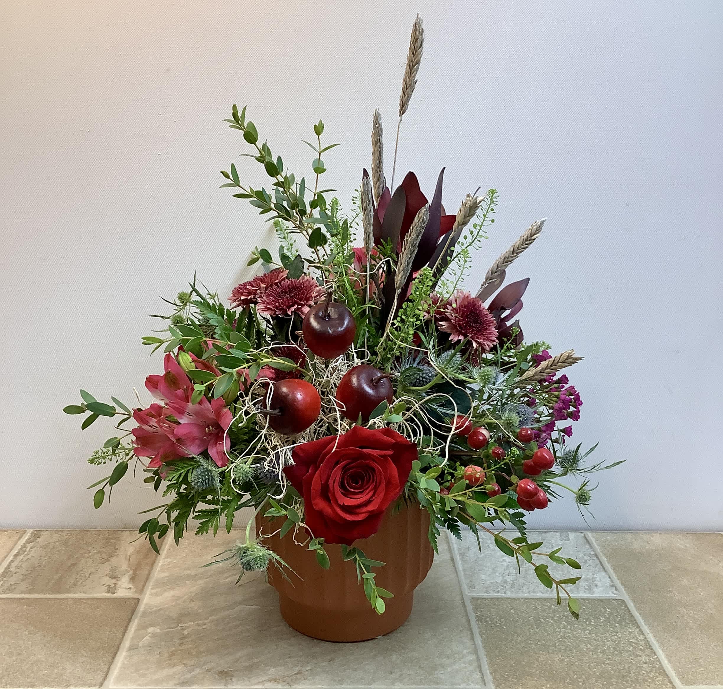 Red rose arrangement in a brown pot with apples and dried wheat