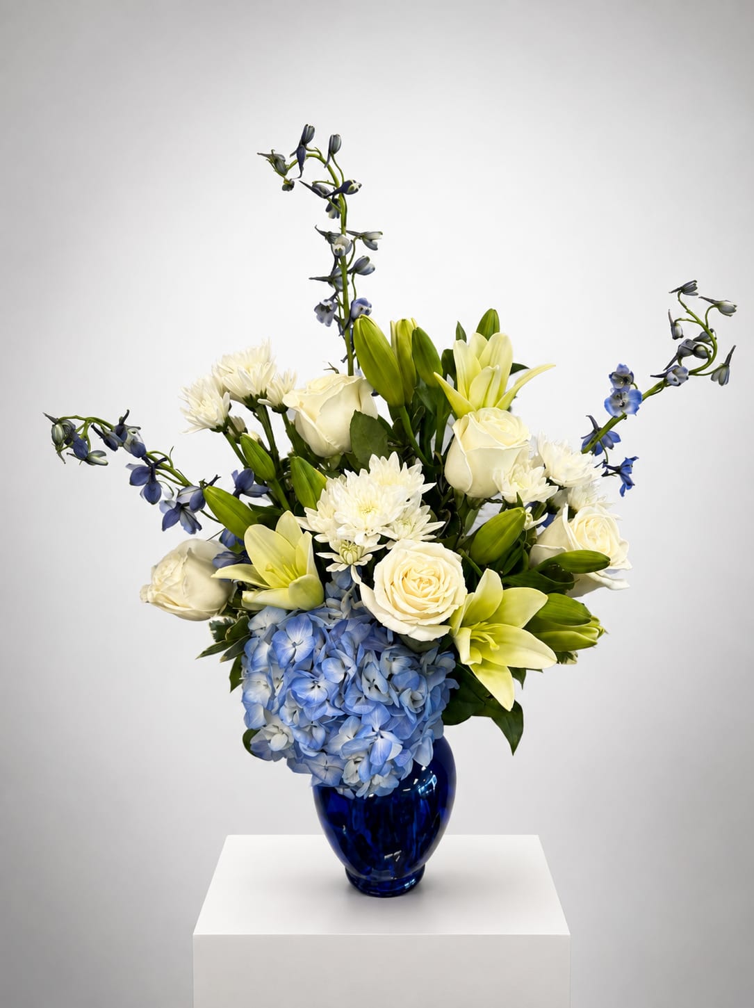 Blue hydrangeas, white roses, lilies, and blue delphinium in a cobalt vase