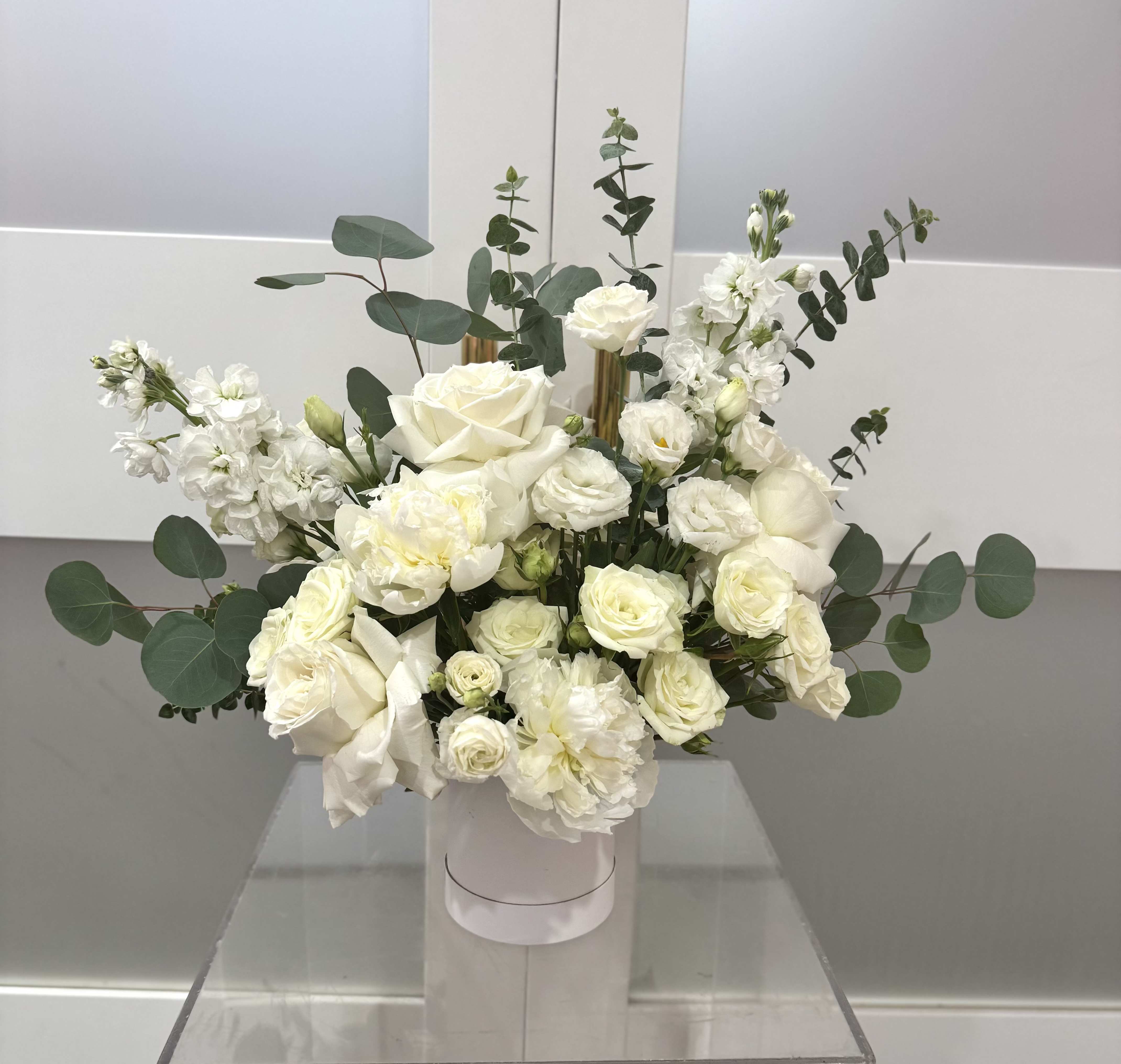 Ivory Grace Luxe Box  - A lush cluster of creamy white roses and delicate lisianthus arranged in a round white hat box creates an elegant statement - perfect as a wedding gift, anniversary surprise, or sympathy arrangement. The Ivory Grace Luxe Box reads soft and refined at first glance, the all-white palette and airy eucalyptus that extend the silhouette into a loose, garden-style bouquet the recipient will feel wrapped in.  This arrangement pairs full-headed roses with ruffled stock and lisianthus for layered texture and gentle fragrance. Presented in a crisp white hat box, the composition stays compact yet abundant, with soft green eucalyptus sprigs adding movement and contrast. The shape is loose and natural rather than tightly formal, giving it a casually luxurious look.  Give this to someone who deserves to feel seen - the bride, a partner marking an anniversary, or a friend needing comfort - and they'll receive a calming, sophisticated bouquet that reads both celebratory and tender. The hat box presentation makes it easy to display on a table or mantle; light care (fresh water source at delivery and out of direct sun) will help blooms stay lovely for several days.
