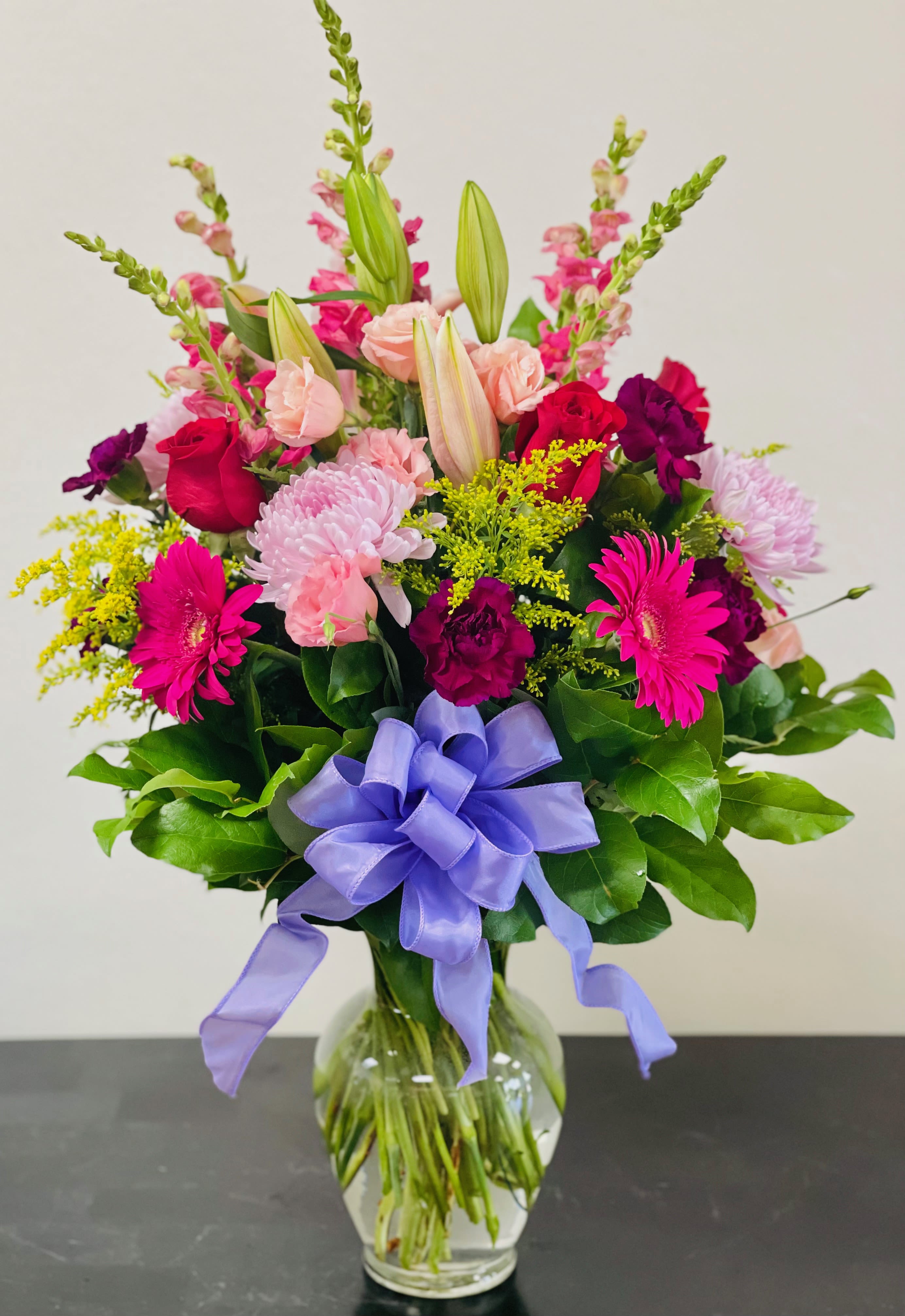Mixed bouquet of pink, purple, and yellow flowers in a glass vase with a lavender ribbon.