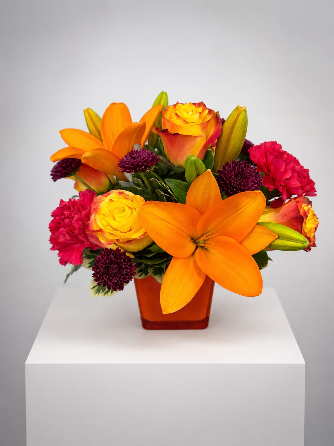 Orange lilies, yellow roses, and pink carnations in an amber vase