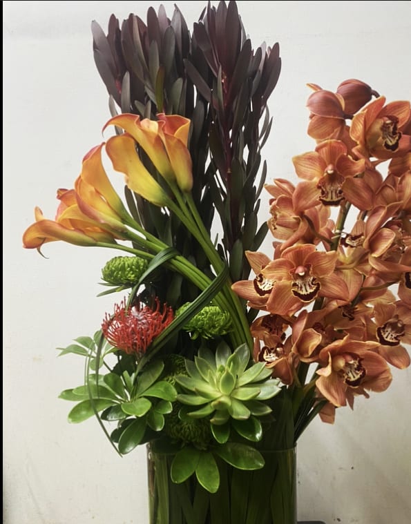 Tall arrangement of orange calla lilies and orange orchids in a glass vase
