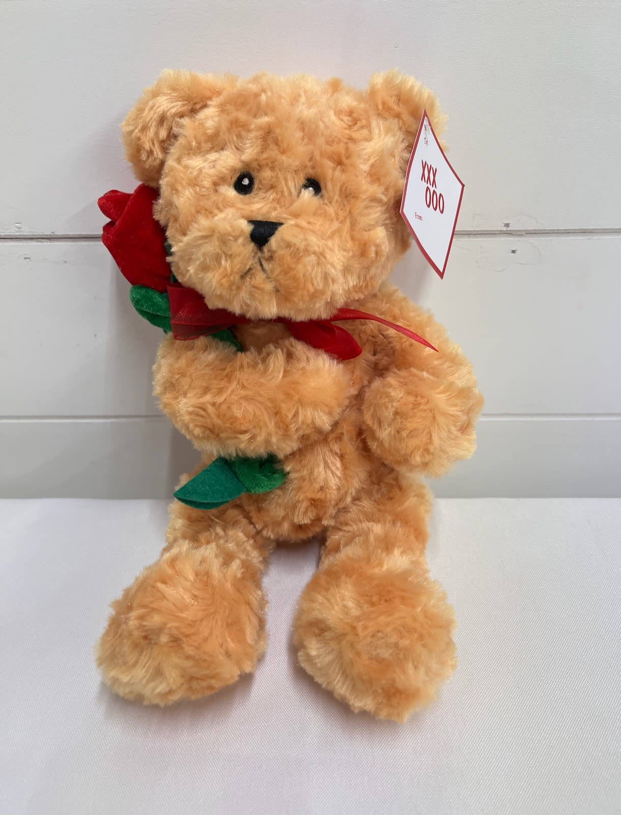 Tan teddy bear holding a red fabric rose with a tag on its ear