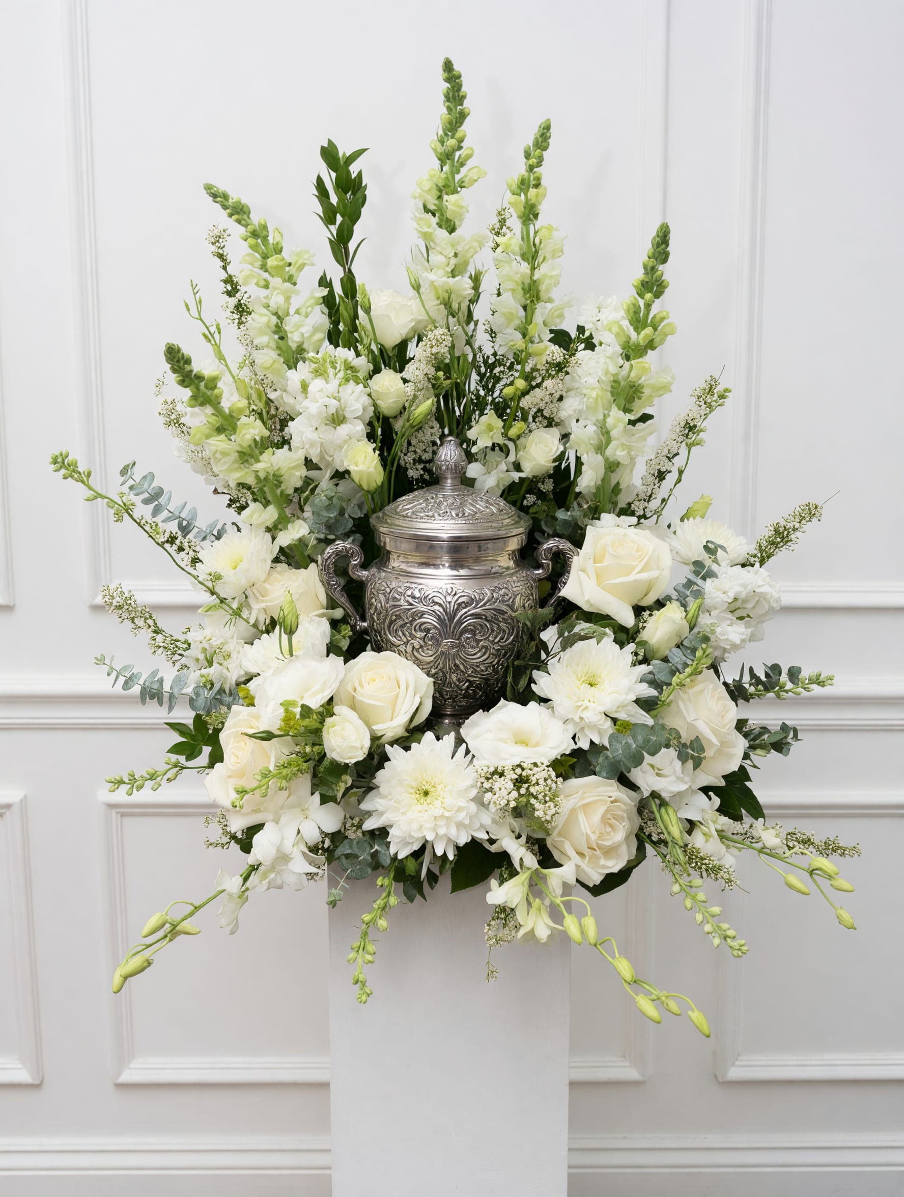 White Garden Tribute - An elegant white floral arrangement designed to surround and honor a loved one’s urn. Created with seasonal white blooms and natural garden movement. Decorative urn shown for display.