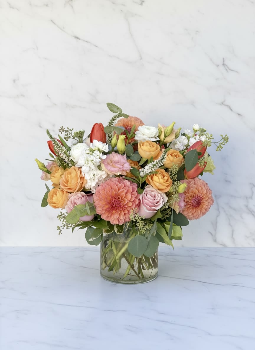 Bay Breeze - Soft peach and pink roses mingle with coral dahlias and delicate white blooms in a garden-style bouquet made to brighten an anniversary or birthday. This Bay Breeze flower arrangement combines romantic tones with lively red tulip accents to create a cheerful, fresh look that's perfect for summer celebrations, saying thank you, or simply thinking of you.  The full, rounded composition features dahlias, roses, tulips, stock, lisianthus, and airy filler flowers arranged in a clear glass vase for a classic, see-through presentation. Soft pastels-peach, pink, and white-are balanced by pops of red and coral, giving the design depth and a layered, textured feel that reads beautifully from every angle.  Send this when you want the recipient to feel truly seen-an uplifting choice for birthdays, new baby congratulations, get-well wishes, or a spontaneous "I'm thinking of you." The arrangement arrives ready to display in its vase; trim stems and refresh the water every few days to keep blooms looking their best.