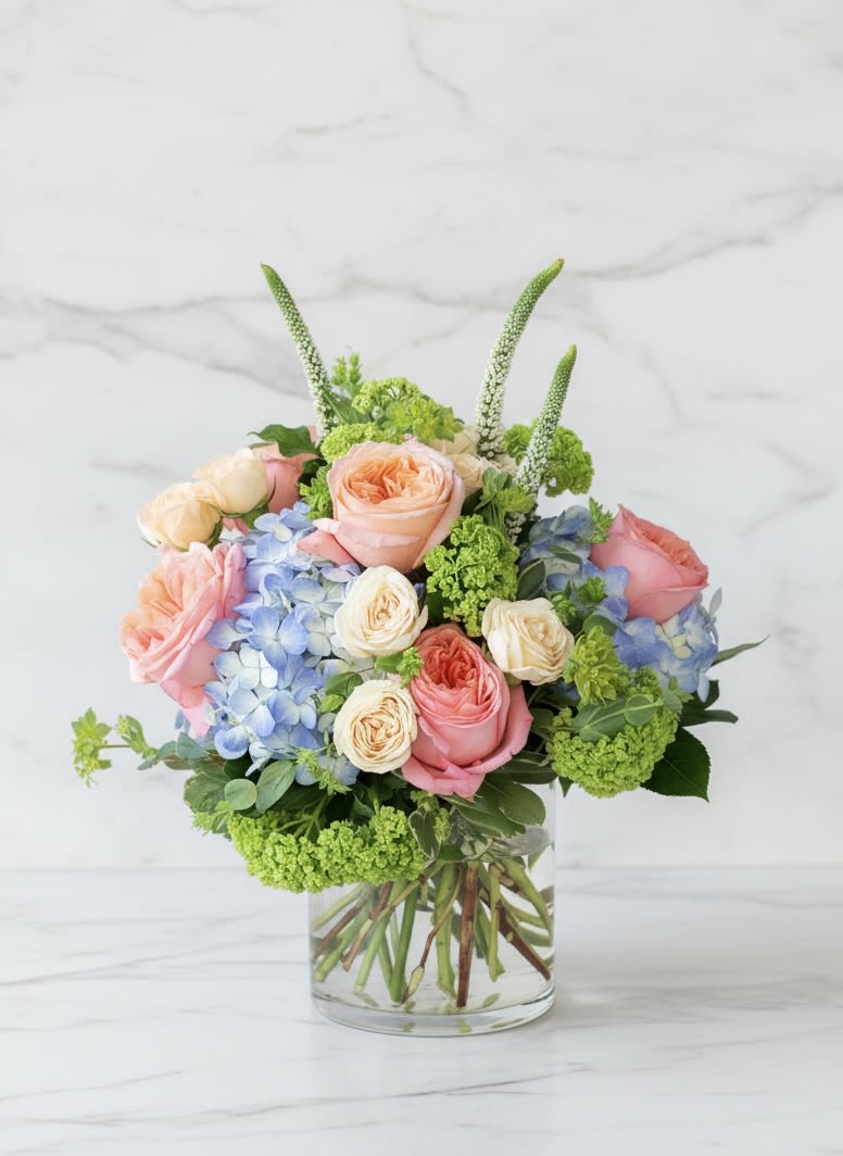 Low arrangement of peach and pink roses, cream roses, and blue hydrangeas in a clear glass vase.