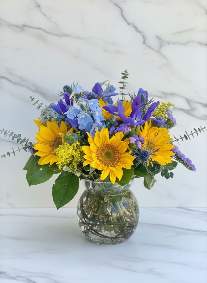 Sun and Sky - Arranged in a bubble bowl, this serene sun and sky design features fresh cut sunflowers nestled amidst a lush collection of blue thistle, iris, and belladonna and is accented with yellow solidago and eucalyptus.