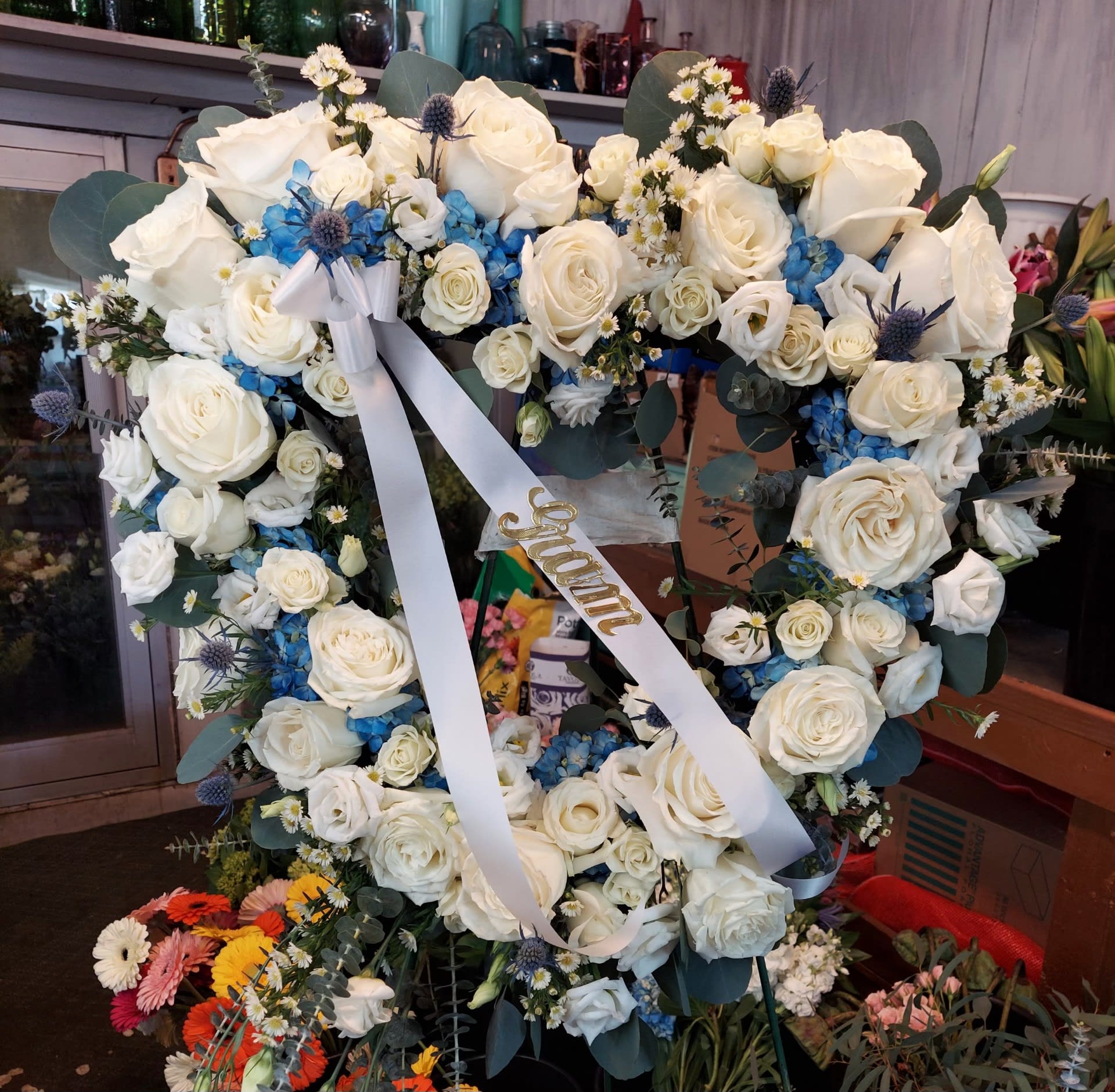 Heart-shaped wreath of white roses with blue flowers and a white ribbon