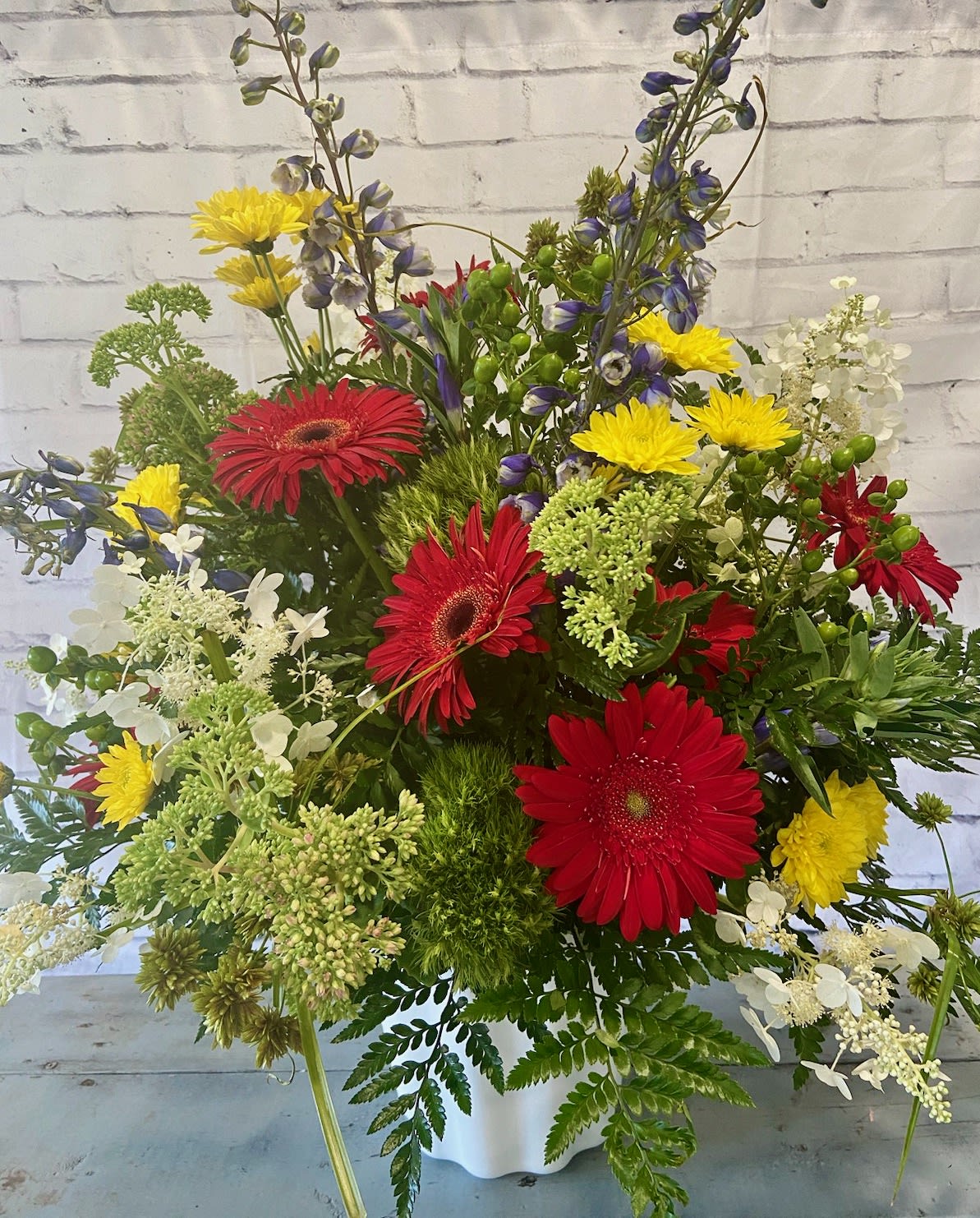Mixed bouquet with red gerbera daisies, yellow blooms, and blue flowers in a white container