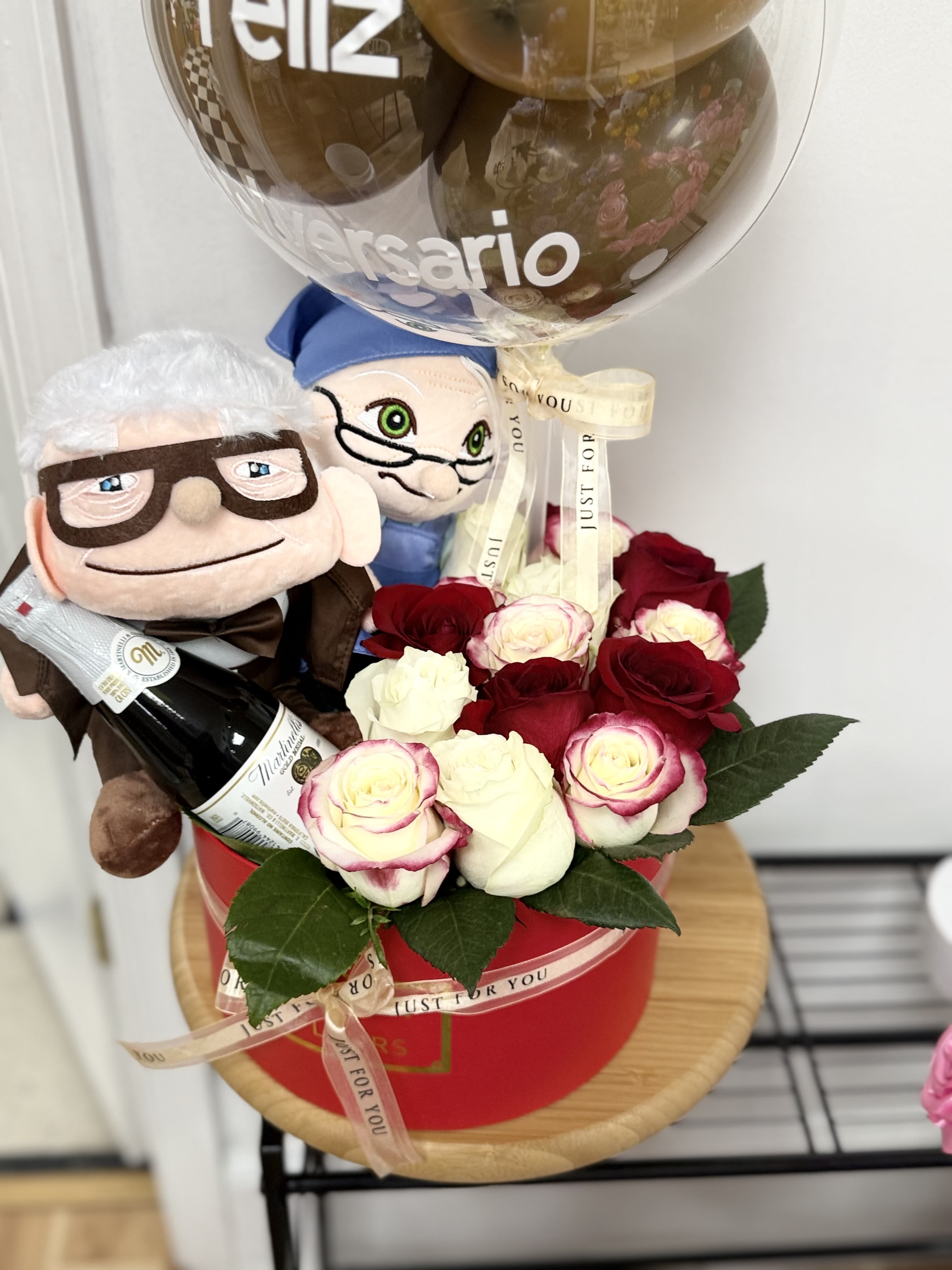 Gift set with red and white roses in a red box, two plush dolls, a bottle, and a clear anniversary balloon.