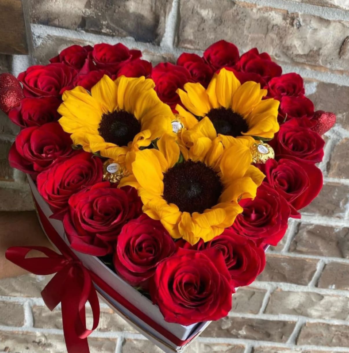 Heart-shaped box filled with red roses, three yellow sunflowers, and gold-wrapped chocolates.