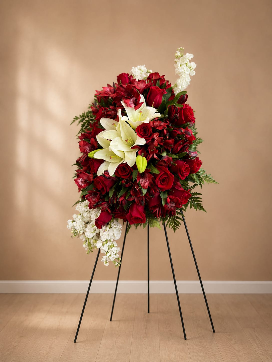 You Care Standing Spray - The spray features stock, roses, lilies and alstroemeria arranged in a layered, formal composition with white cascading blooms that trail from the lower left and upper right for graceful movement. Mounted on a black metal easel for a classic ceremonial presentation, the piece reads tall and substantial at services or graveside. Care: keep flowers in a cool place before use and allow florists' mechanics to hydrate the foam until delivery/setup at the service.