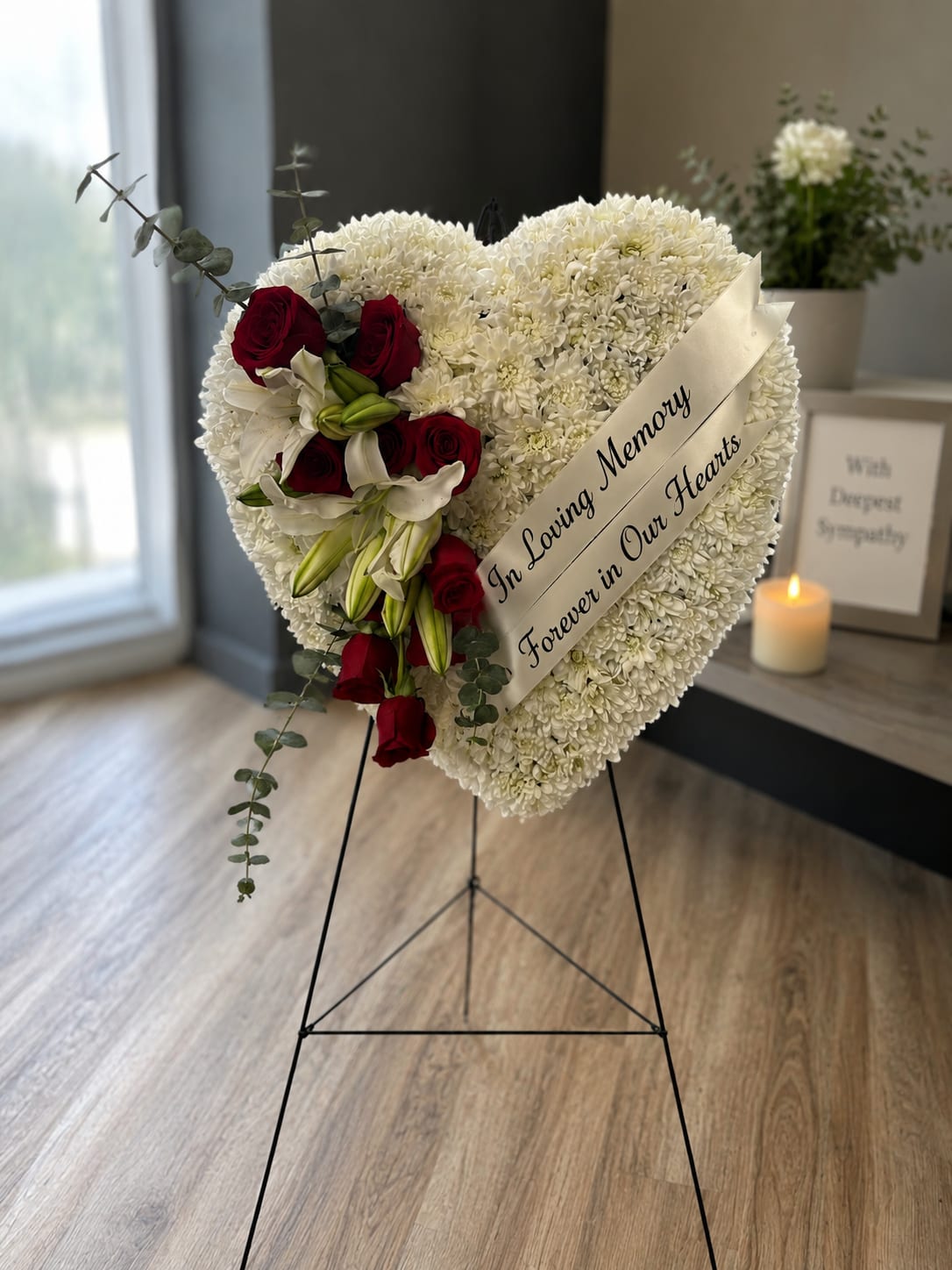 Pure Love Standing Spray - A heart-shaped sympathy standing spray that reads as a quiet, dignified tribute-tightly packed white chrysanthemums form the canvas while a diagonal cluster of deep red roses and white lilies creates a striking, heartfelt focal point for a funeral or memorial service. The satin sympathy ribbon printed with "In Loving Memory" and "Forever in Our Hearts" reinforces the message of remembrance, so the person you're honoring will feel truly remembered and respected.  Crafted for a formal setting, the arrangement mounts on a black metal easel stand and includes a small candle to add a gentle, contemplative touch. The mix of chrysanthemum texture, velvety rose petals, and sculpted lily blooms provides visual depth and a comforting palette of white, red, and green. Sized to stand visibly beside a casket or memorial table, it arrives ready to display and offers enduring sympathy without fuss-simple care is keeping it cool and away from direct sun to maintain freshness during services.