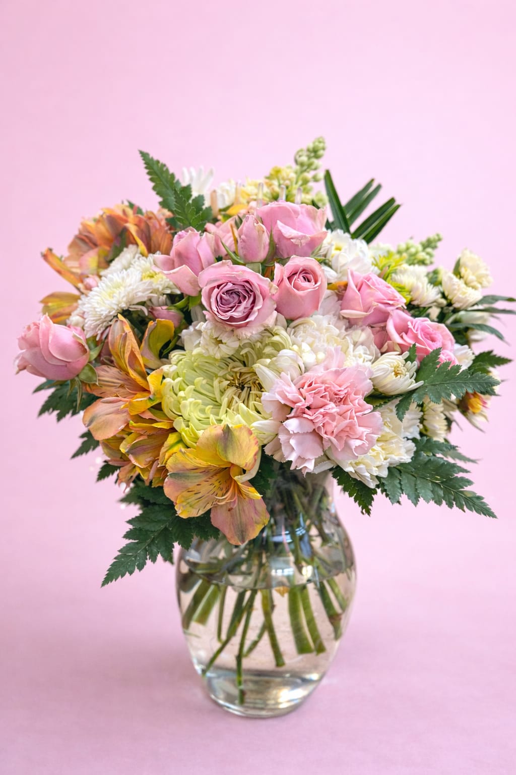 Forever Unique Bouquet - A lush, garden-style arrangement that feels both romantic and cheerful-perfect as a birthday bouquet or anniversary flowers to make someone feel truly celebrated. Soft pink roses and pale carnations take center stage while warm peach-and-yellow alstroemeria and crisp white chrysanthemums add contrast and brightness, creating a pastel palette that reads like spring in bloom.  This mixed-color arrangement is densely layered for an abundant, rounded silhouette and is presented in a clear glass vase that showcases the fresh green stems and soft yellow-green hydrangea clusters. The combination of textures-from the velvety roses to the ruffled carnations and full hydrangea heads-gives the bouquet a hand-arranged, freshly picked look.  Choose this arrangement when you want to say "you're thought of" with warmth and joy-ideal for mothers, partners, close friends, or anyone who loves soft pastels and a lush presentation. Vase dimensions and exact stem counts vary by size; stems arrive hydrated and should be refreshed with clean water every 2-3 days and trimmed at an angle for longest enjoyment.
