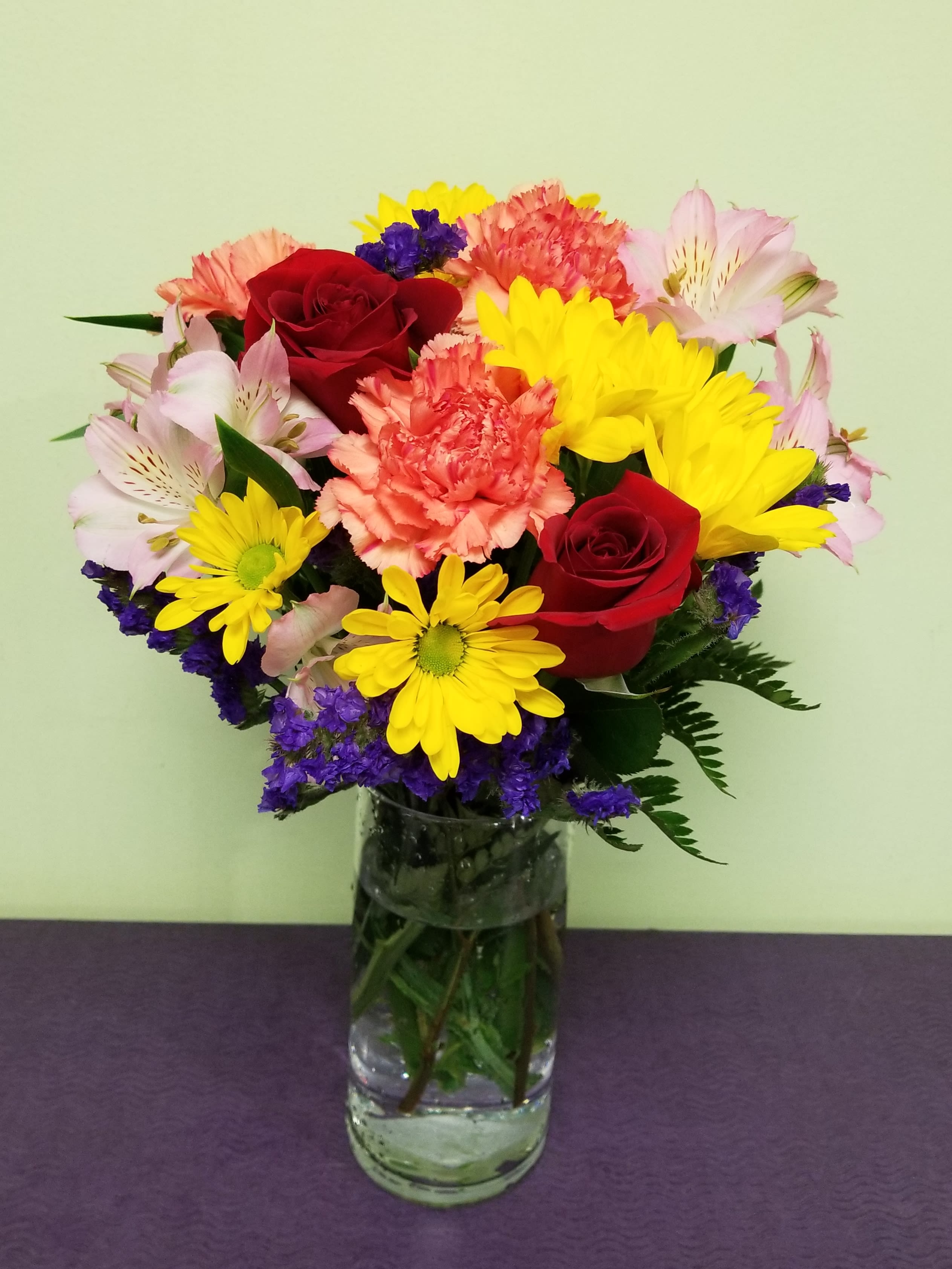 Mixed bouquet of red roses, yellow daisies, pink alstroemeria, and carnations in a glass vase