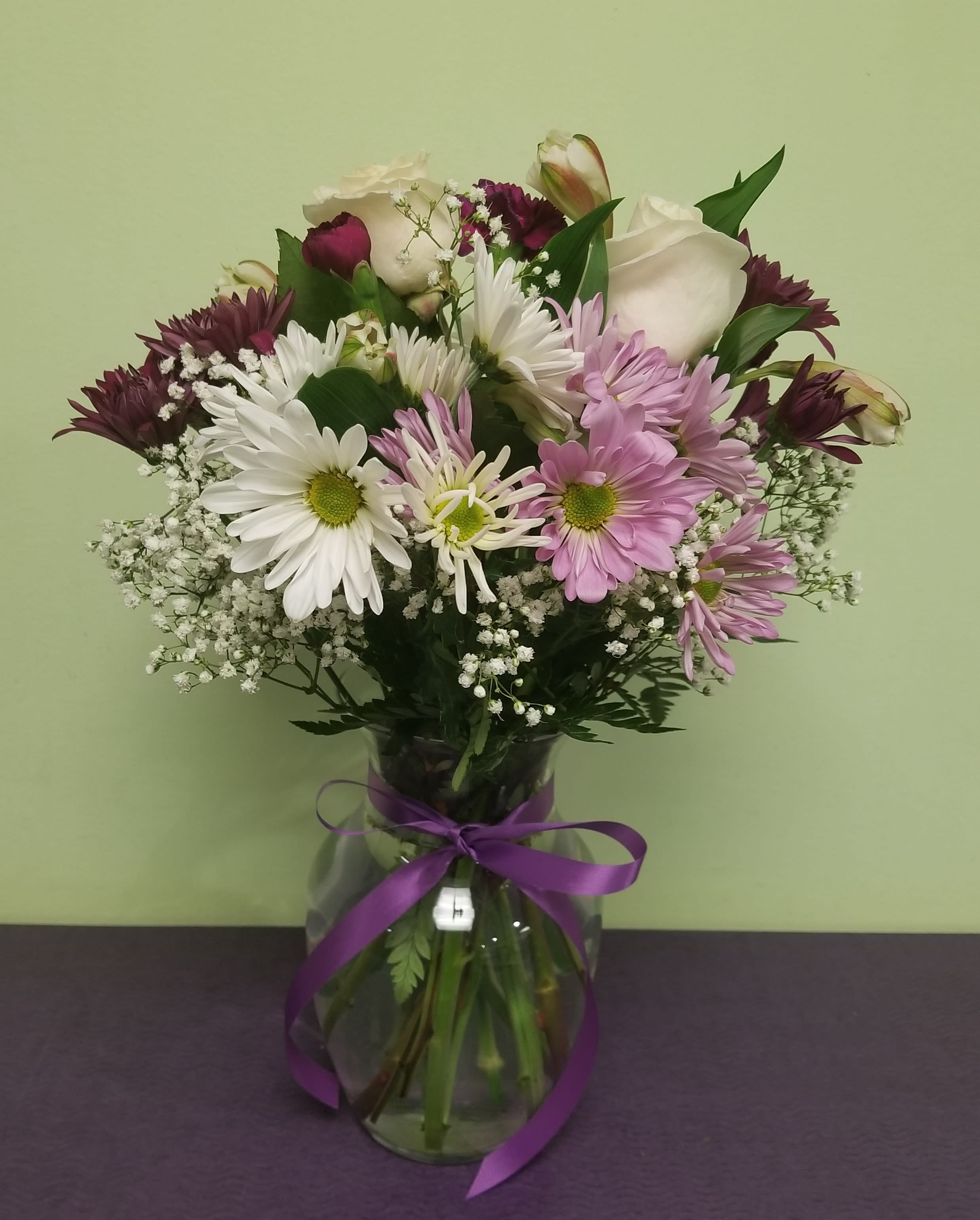 Mixed bouquet of daisies, roses, and purple blooms in a glass vase with ribbon